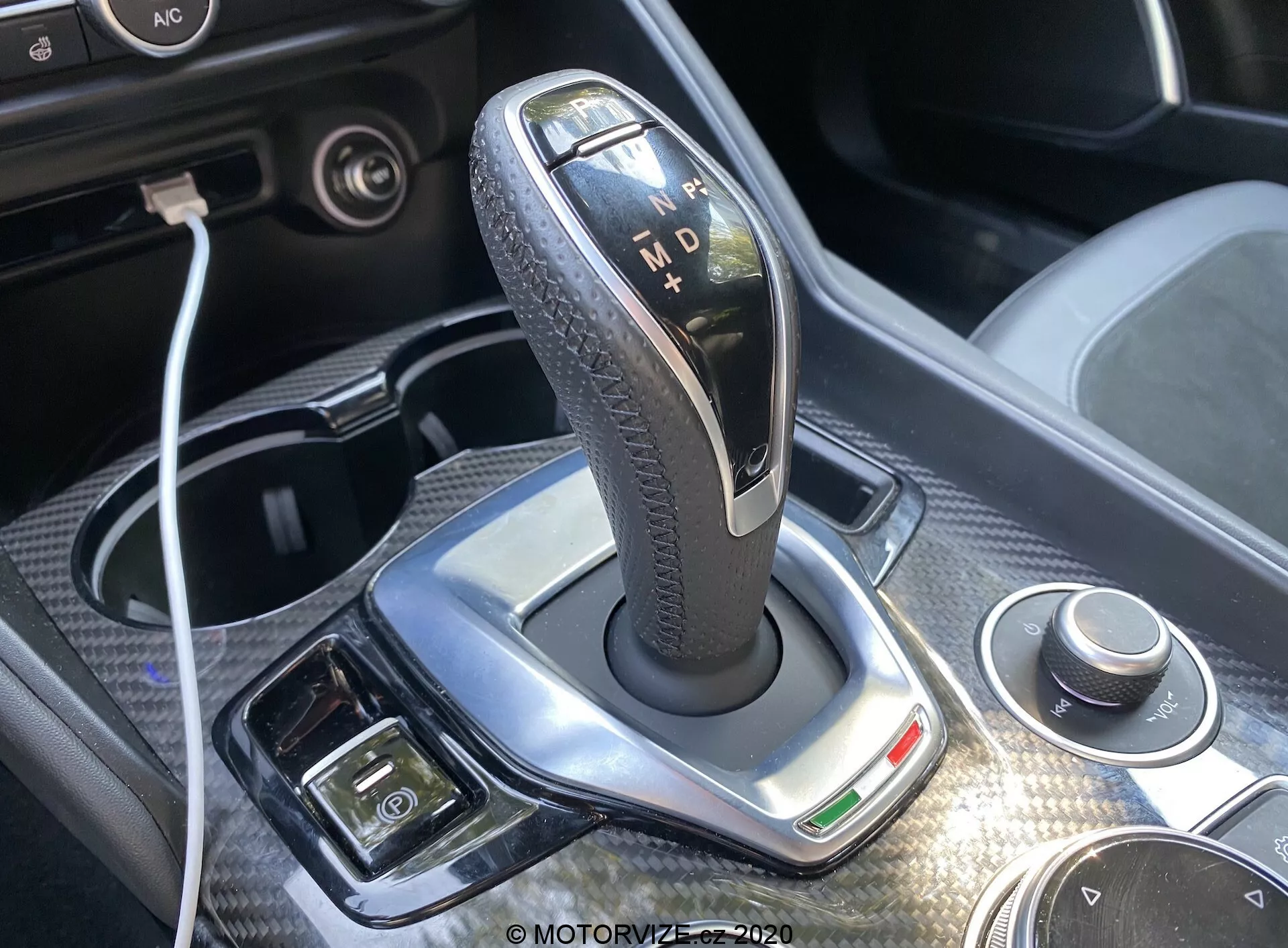 Pogled od blizu na prestavno ročico samodejnega menjalnika Alfa-Romeo Giulia 2016 s elegantnim poliranim srebrnim robom, črnim usnjenim teksturiranim ročajem in osvetljenimi indikatorji za 'P', 'R', 'N', 'D' in 'M'. Svetla dnevna svetloba poudarja dizajn, v bližini pa so vidni vrtljiv gumb in USB povezava. Prisotna je tudi kromirana obloga, zelena indikatorska lučka in dodatni gumbi za elektronsko parkirno zavoro in klimatski sistem, kar ponuja vpogled v luksuz in napredno tehnologijo notranjosti avtomobila.
