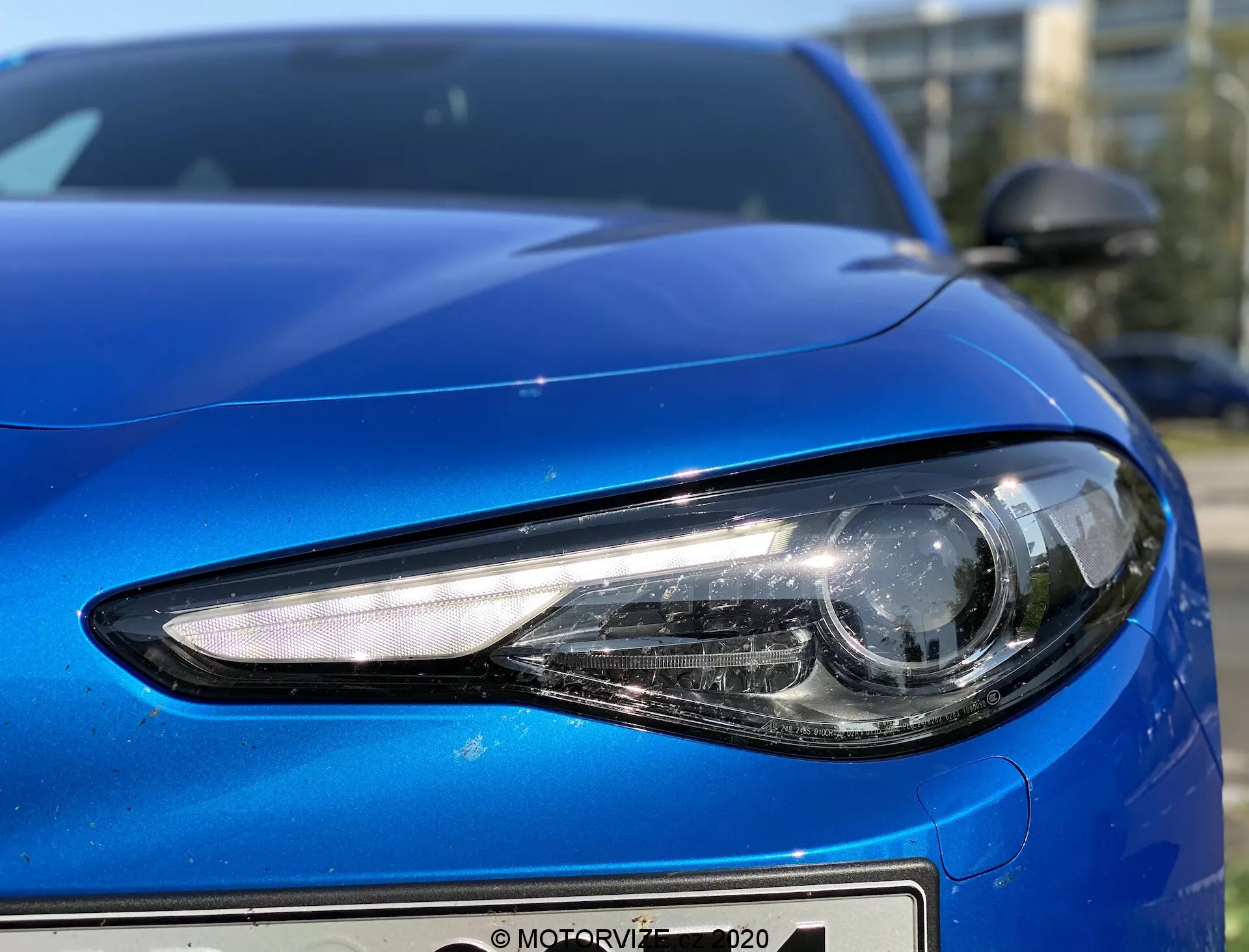 Pogled od blizu levo stran modrega vozila Alfa Romeo Giulia 2016, posnet pod nizkim kotom, ki prikazuje podrobnosti žarometa in krivulje zasnove avtomobila. Sodobni žaromet vključuje LED ali HID osvetlitev z elegantno, kotno obliko in glavni snop v projektorskem slogu za usmerjeno svetlobo. Pokrov motorja ima gladek, konturiran dizajn z odsevnim kovinskim zaključkom, sprednji odbijač pa ima minimalistično zasnovo. Stransko ogledalo je v barvi karoserije in aerodinamične oblike, delno je vidno v zgornjem desnem kotu, kar dopolnjuje aerodinamični profil avtomobila.