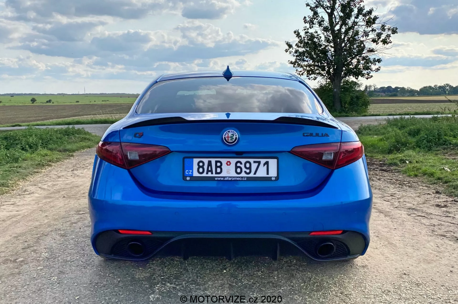 Zadnji pogled na svetlo modri Alfa Romeo Giulia 2016 limuzino, posnet iz perspektive oči. Slika prikazuje celoten zadnji del avtomobila, centriran v okvirju, s jasno vidno registrsko tablico, zadnjimi lučmi in logotipom proizvajalca. Značilnosti vključujejo dvosmerni izpušni sistem, elegantne moderne zadnje luči, difuzorski element na odbijaču, spojler na prtljažnikih vratih, parkirne senzorje in zadnji brisalec, kar nakazuje ravnovesje med športnim značajem, varnostjo in praktičnostjo. Okolje je podeželsko območje s plosko pokrajino in delno oblačnim dnevnim nebom.