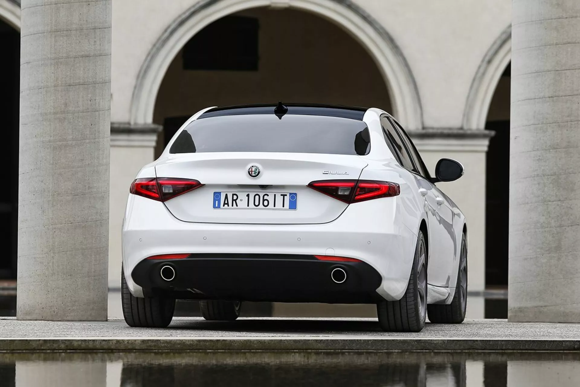 Bela Alfa Romeo Giulia letnik 2016, fotografirana iz zadnjega tričetrtinskega kota, ki poudarja njen športni zadnji profil, značilne zadnje luči, dvojne izpušne cevi in ​​elegantne oblikovalske značilnosti na ozadju evropske arhitekture