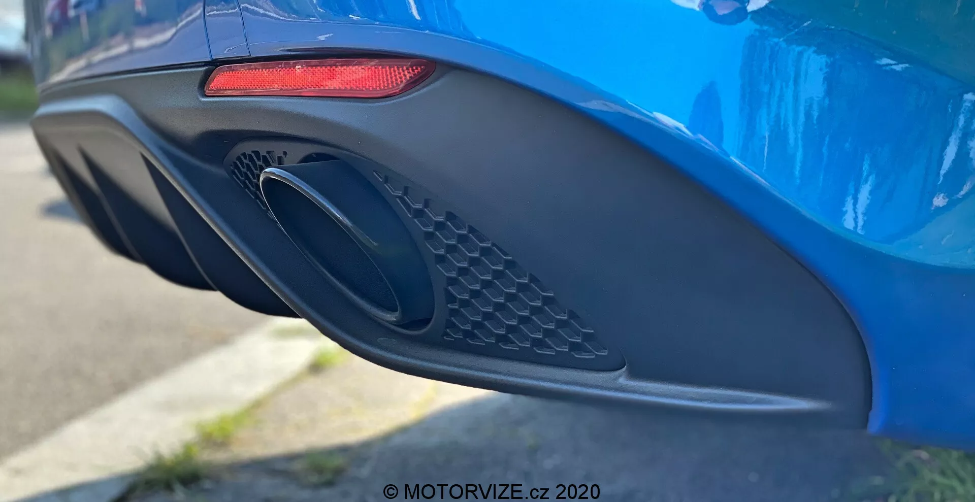 Od blizu stranski pogled na zadnji odbijač in izpušni del modrega Alfa Romeo Giulia 2016, ki prikazuje sodobne oblikovne značilnosti, vključno z raztegnjeno vodoravno rdečo LED zadnjo lučjo, velikim ovalnim izpušnim odprtino z mrežastimi podrobnostmi in aerodinamično oblikovanim odbijačem z različnimi krivinami in vdolbinami, fotografirano z majhno globino polja za poudarjanje detajlov avtomobila.