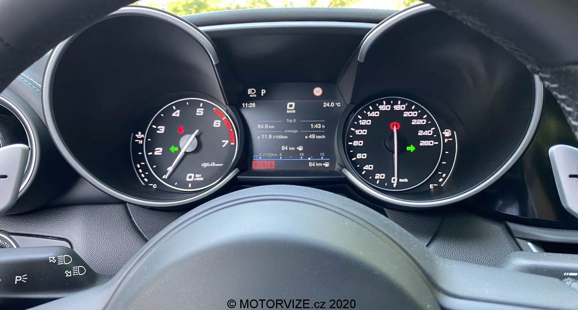 Pogled voznika na instrumentno ploščo Alfa Romeo Giulia 2016 z osrednjim digitalnim zaslonom, ki prikazuje hitrost, prestavo, temperaturo in prikaz prevoženih kilometrov, obdanim z analognim merilnikom hitrosti in merilnikom vrtljajev, ki prikazuje mešanico tradicionalnega oblikovanja in sodobne tehnologije, z delnim pogledom na krmilne elemente za avdio in tempomat na volanu.