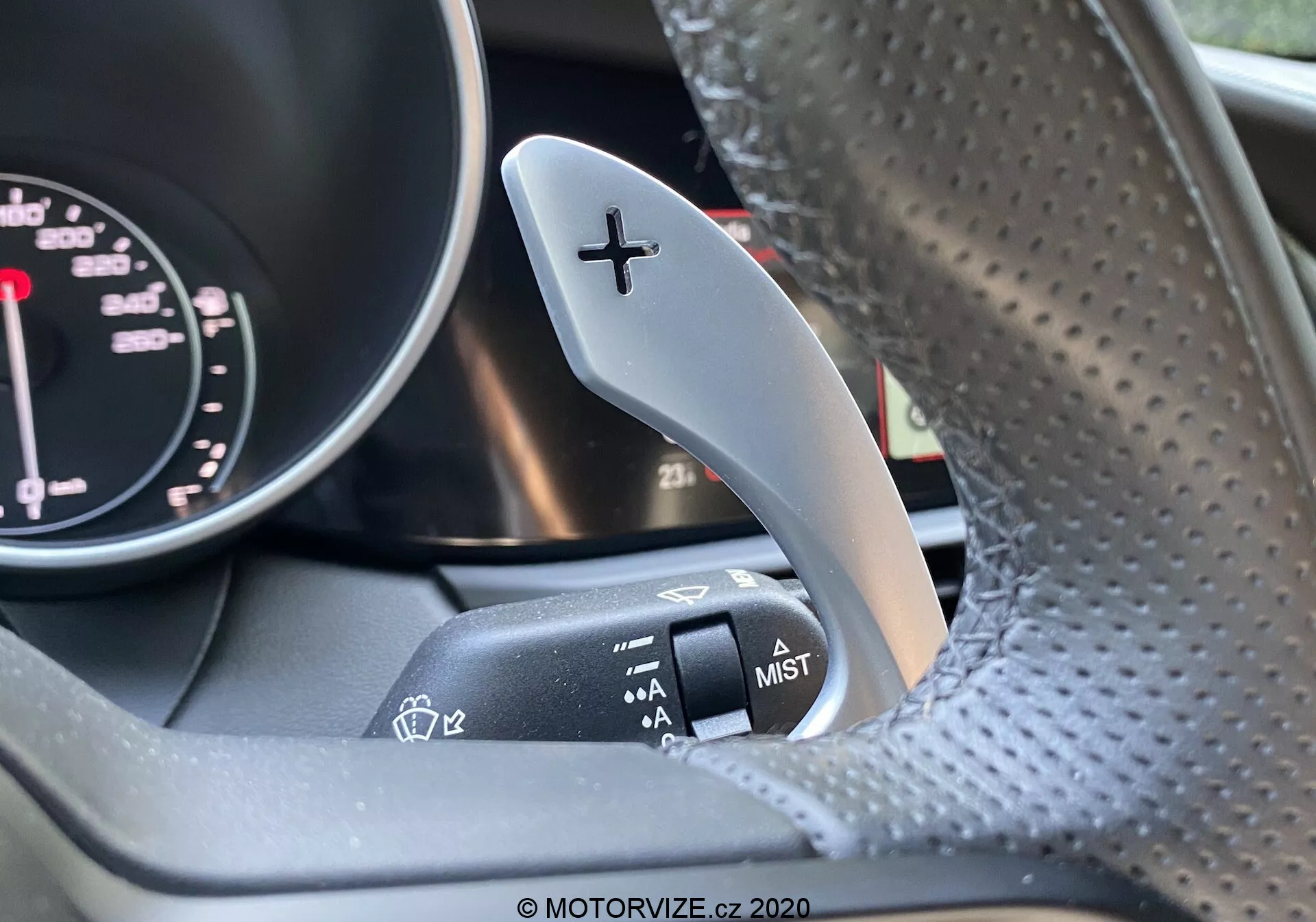 Posnetek notranjosti Alfa Romeo Giulia 2016, osredotočen na desno prestavno ročico z oznako '+' za prestavljanje navzgor in sosednjo ročico za nadzor brisalcev na volanskem drogu. Del tahometra iz instrumentne plošče je rahlo viden. Volan iz perforiranega usnja je rahlo zamegljen. Slika poudarja kontrast med kovinsko prestavno ročico in matiranim zaključkom ročice za nadzor brisalcev.