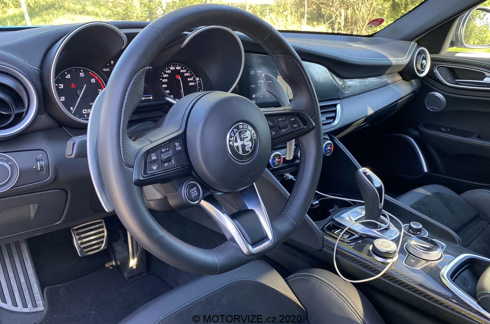 Notranjost avtomobila Alfa Romeo Giulia 2016, osredotočena na voznikovo stran iz perspektive sovoznika, prikaz volana s tremi kraki z večfunkcijskimi gumbi, vidnim logotipom proizvajalca, instrumentno ploščo z analognimi številčnicami in digitalnim zaslonom, sredinsko konzolo z gumbi za nadzor zvoka in klimatske naprave, ročno prestavno ročico z usnjeno prevleko, izbirnik načinov vožnje, sprednje sedeže s temnim oblazinjenjem in delno vidno voznikovo vratno ploščo z gumbi za okna in ogledala.