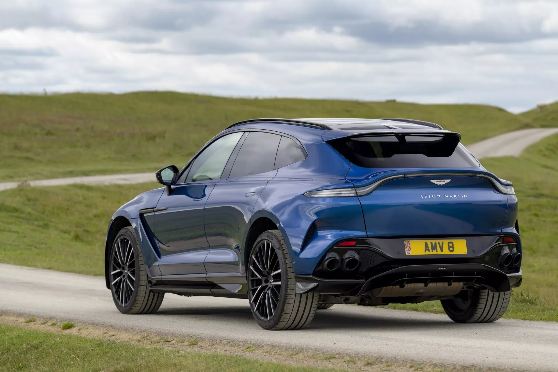 Luksuzni crossover SUV Aston Martin DBX 2020 v modri barvi, prikazuje tričetrtinski zadnji in stranski profil s športnim dizajnom, vključno z metalnim zaključkom, zadnjim spojlerjem, vitkimi zadnjimi lučmi, zlitimi platišči in možnimi funkcijami visoke tehnologije.