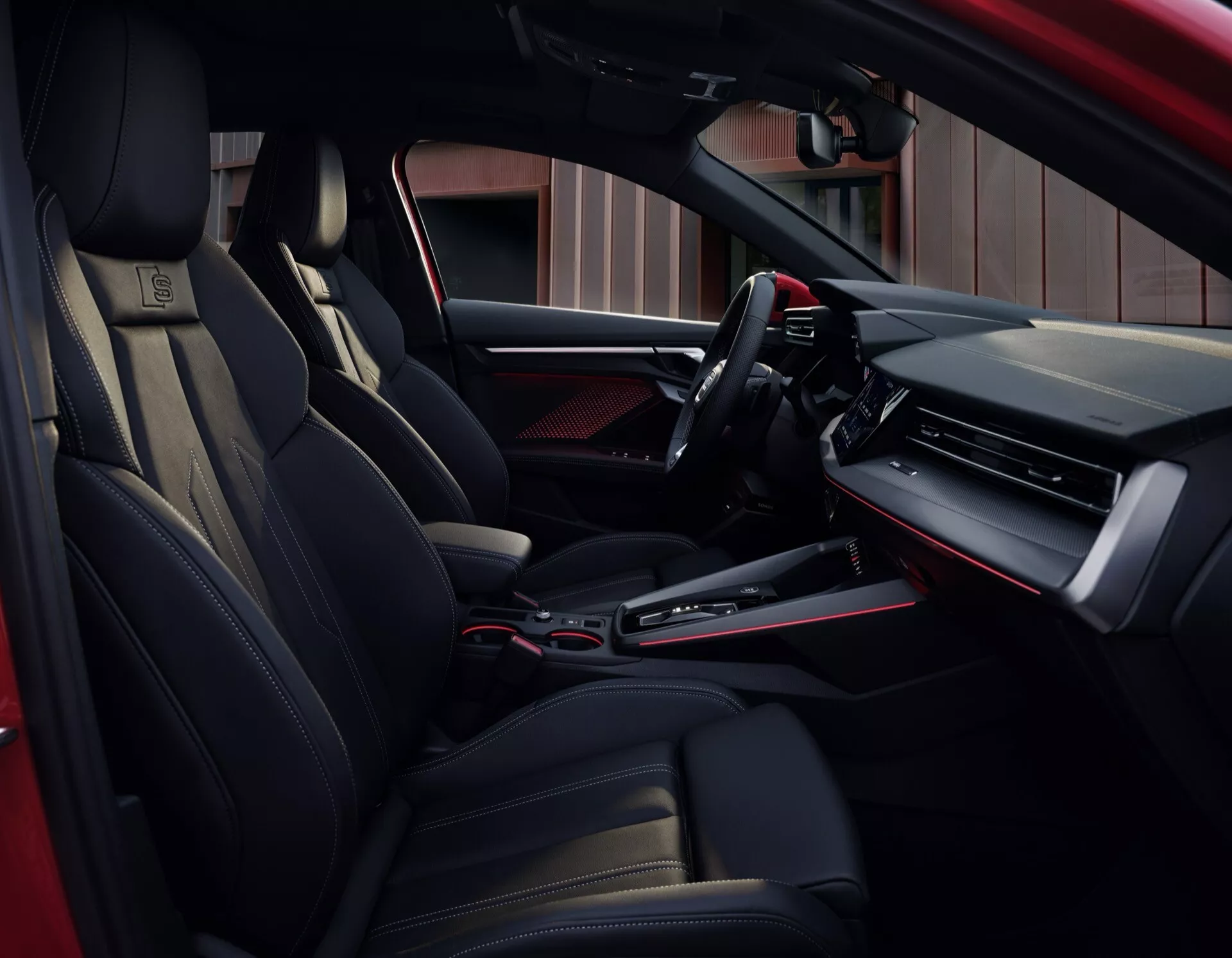 Notranji pogled na Audi A3 2024 (8Y-facelift) s poudarkom na voznikovi strani iz perspektive zadnjega potnika, prikazuje elegantno armaturno ploščo z digitalnim zaslonom in velikim zaslonom za infotainment, multifunkcijskim volanom z naprednimi kontrolnimi funkcijami, športnimi ergonomskimi sedeži s prevleko iz usnja, avtomatsko prestavno ročico na sredinski konzoli in luksuznimi črnimi ter kovinskimi poudarki po celotni kabini.