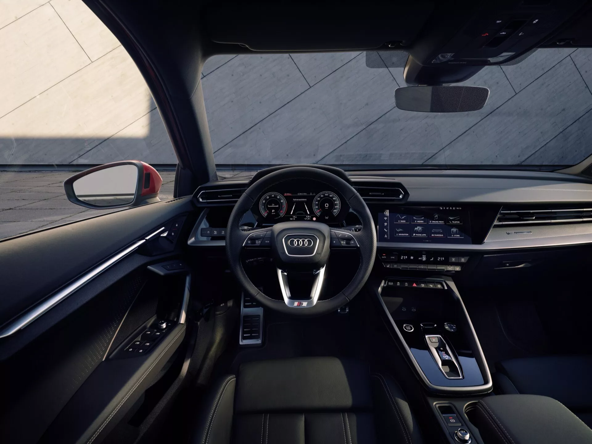 Notranji pogled na Audi A3 2024 (8Y-Facelift), ki prikazuje perspektivo voznika z zadnje klopi, s športnim volanom z večnamenskimi gumbi, podaljšanim digitalnim armaturnim ploščo in infotainment zaslonom, elegantno sredinsko konzolo z minimalnimi fizičnimi kontrolami, sodobnimi prednjimi sedeži iz premium materialov in s kontrastnim šivanjem ter ambientalno osvetlitvijo.