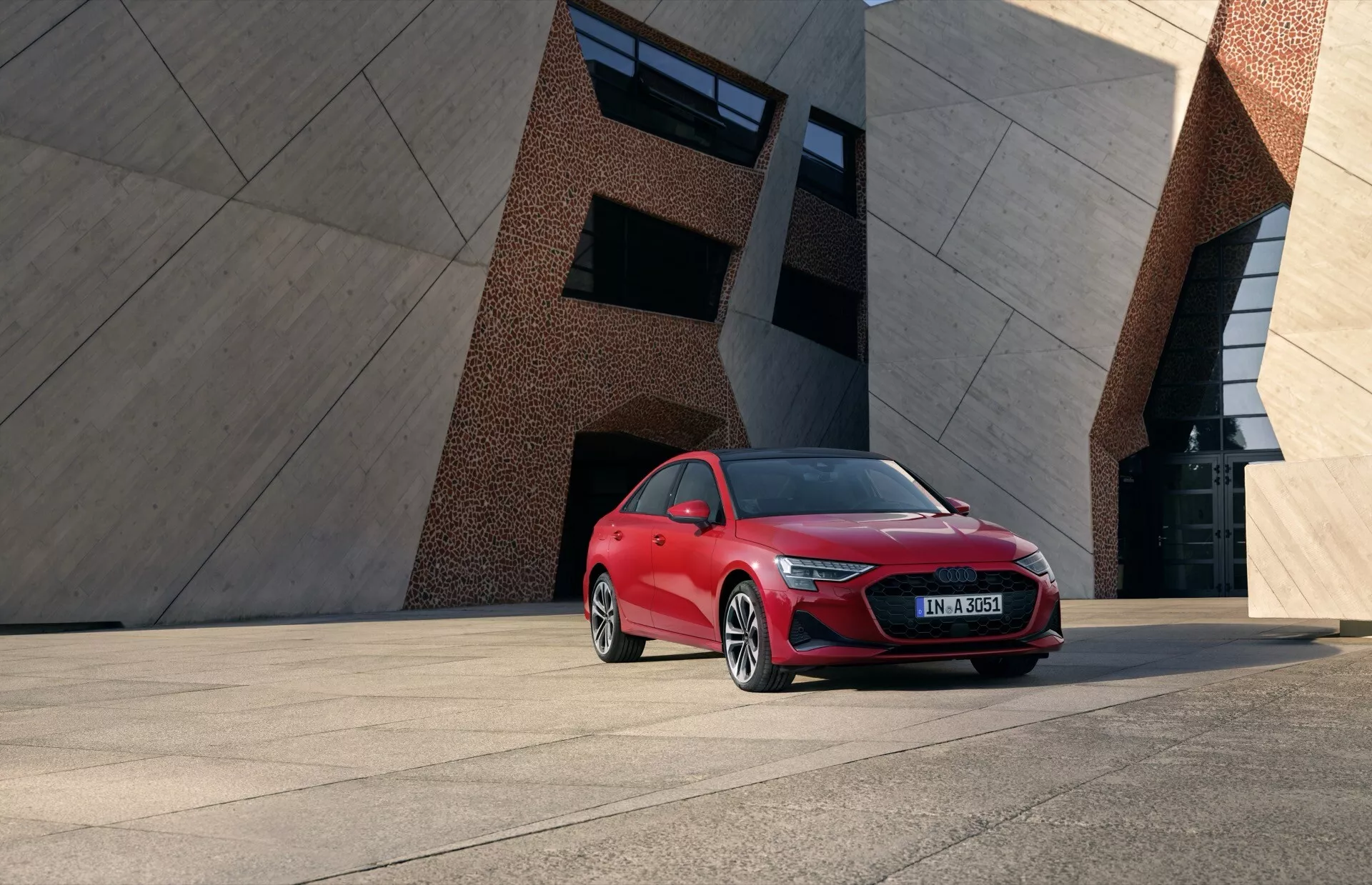 Audi A3 8Y Facelift 2024 v svetlo rdeči barvi, ki prikazuje LED žaromete, izrazito masko in lita kolesa proti sodobnemu urbanemu trgu in geometrični arhitekturi