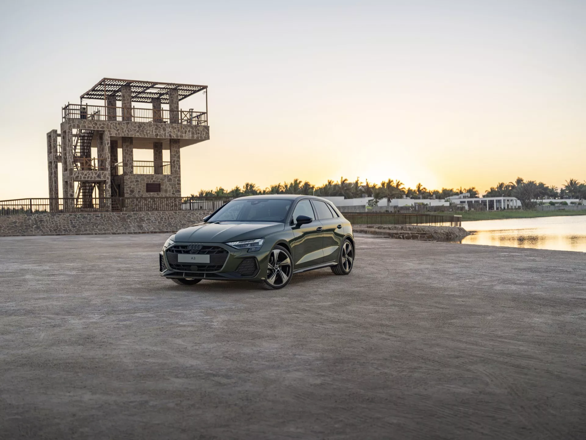 Audi A3 (8Y-Facelift) 2024 postavljen na odprtem tlakovanem območju ob sončnem vzhodu ali zahodu z zgradbo v gradnji in telesom vode v ozadju, prikazuje vozilovo elegantno LED razsvetljavo, kiparsko oblikovanje karoserije in uspešnostno orientirana lita kolesa.