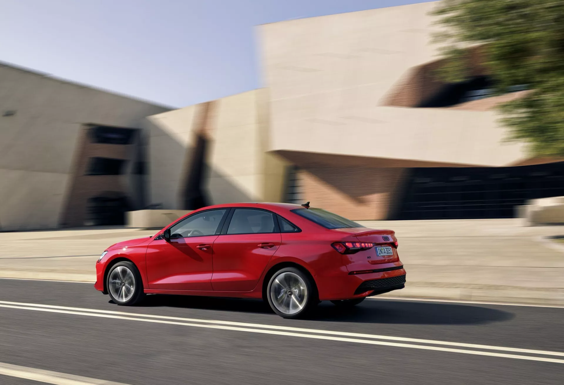 Audi A3 sedan 2024 v gibanju, rdeče barve, posnet s trehčetrtinskega zadnjega vidika, ki prikazuje zlitinska kolesa, aerodinamično obliko karoserije, LED zadnje luči, dvojne izpuhe in učinek zameglitve gibanja na mestni ulici ob zlati uri