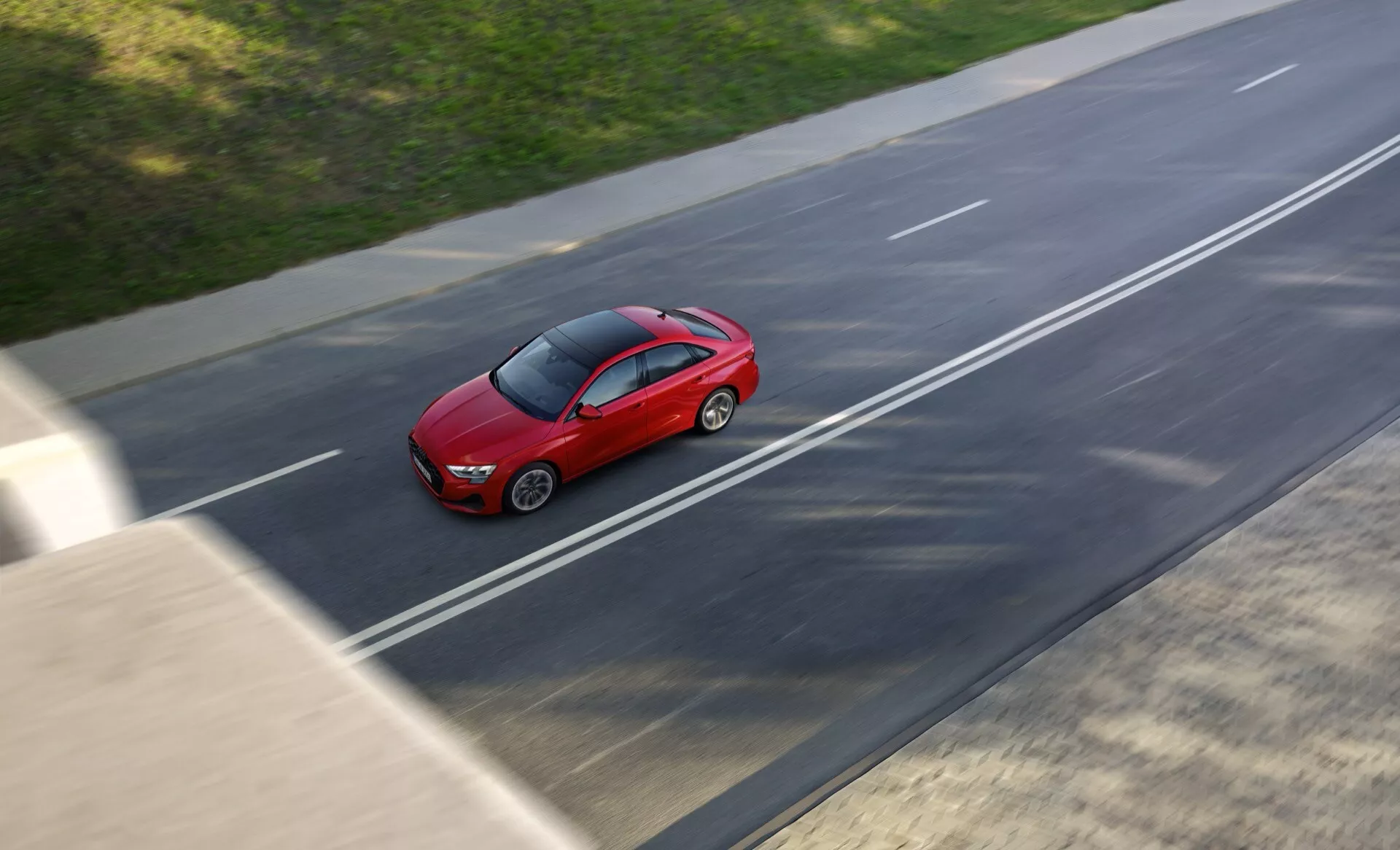 Model Audi A3 8Y facelift 2024 v svetlo rdeči barvi, ujet iz ptičje perspektive med vožnjo po večpasovni avtocesti, prikazuje rahlo zameglitev gibanja in razkazuje elegantno, aerodinamično obliko sedana v sončnih pogojih