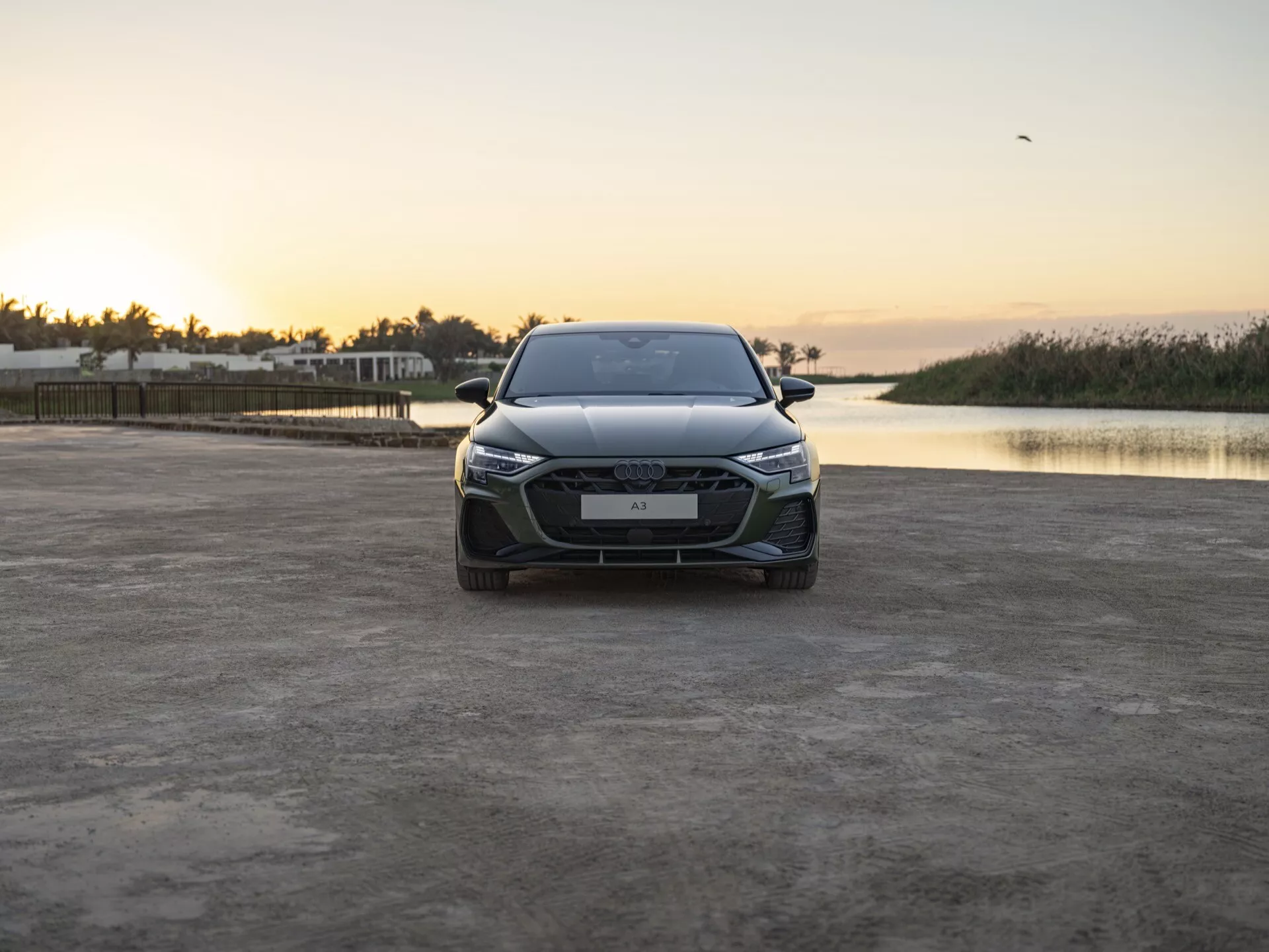 Sprednji pogled na Audi A3 (8Y-Facelift) leto 2024 na nepopravljivi površini z izstopajočo rešetko, LED žarometi z dnevnimi lučmi, naprednimi sistemi pomoči vozniku na odbijaču in oblikovanim pokrovom motorja, med mirnim vzhodom ali zahodom sonca z ozadjem mirne vode in samotnega ptica na nebu, kar poudarja sodobno zasnovo in tehnološke napredke avtomobila proti naravni sceni