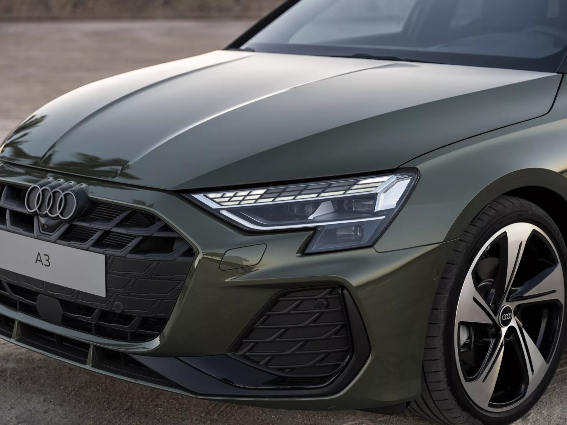 Pogled od blizu na sprednji levi del Audi A3 2024 (8Y-Facelift), ki prikazuje izrazite linije pokrova, levo LED žarometno enoto, šesterokotno sprednjo masko z vodoravnimi letvami in delček uspešnostno naravnanega zlitinskega kolesa in blatnika