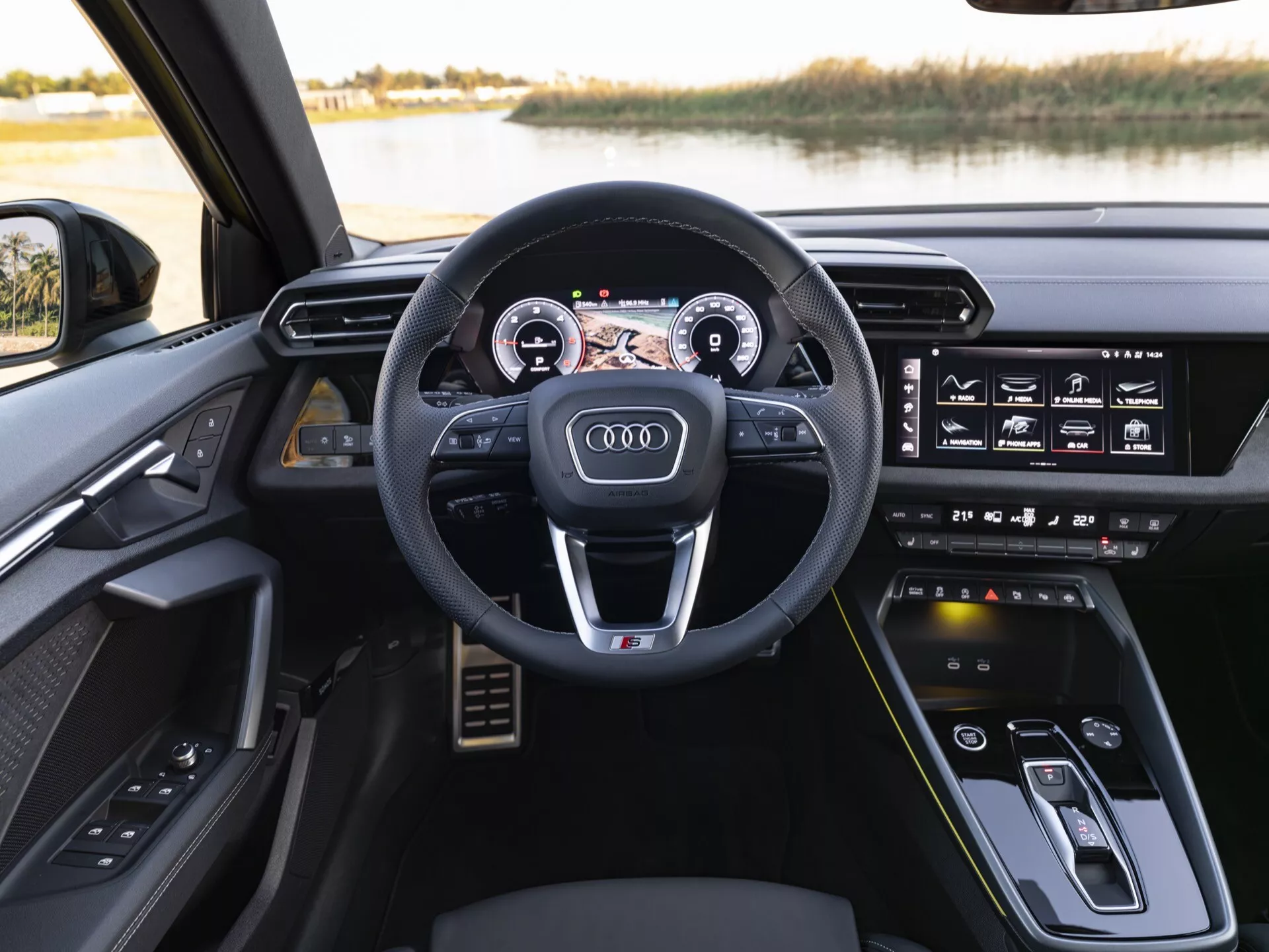 Notranjost Audija A3 (8Y-Facelift) 2024 iz perspektive voznika s športnim multifunkcijskim volanom, ovitim v usnje, povsem digitalnim kokpitom Audi Virtual Cockpit in zaslonom na dotik za informacijsko-zabavni sistem na armaturni plošči, postavljeno proti mirnemu zunanjemu okolju ob jezeru
