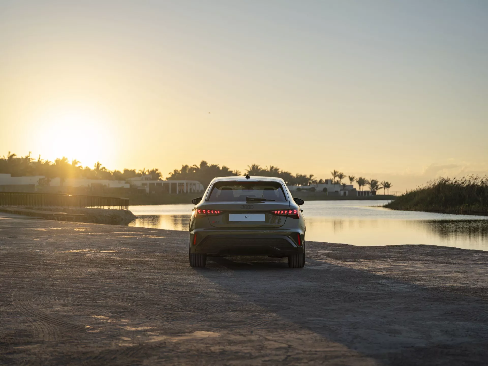 Zadnji pogled na Audi A3 2024 (8Y-Facelift) ob mraku, pozicioniran v središču proti ozadju sončnega zahoda z mirno vodno površino na desni, poudarjajoč njegov sodoben aerodinamični dizajn in edinstveno zadnjo luč, ki sega čez širino vozila na rahlo mokri teksturirani cesti