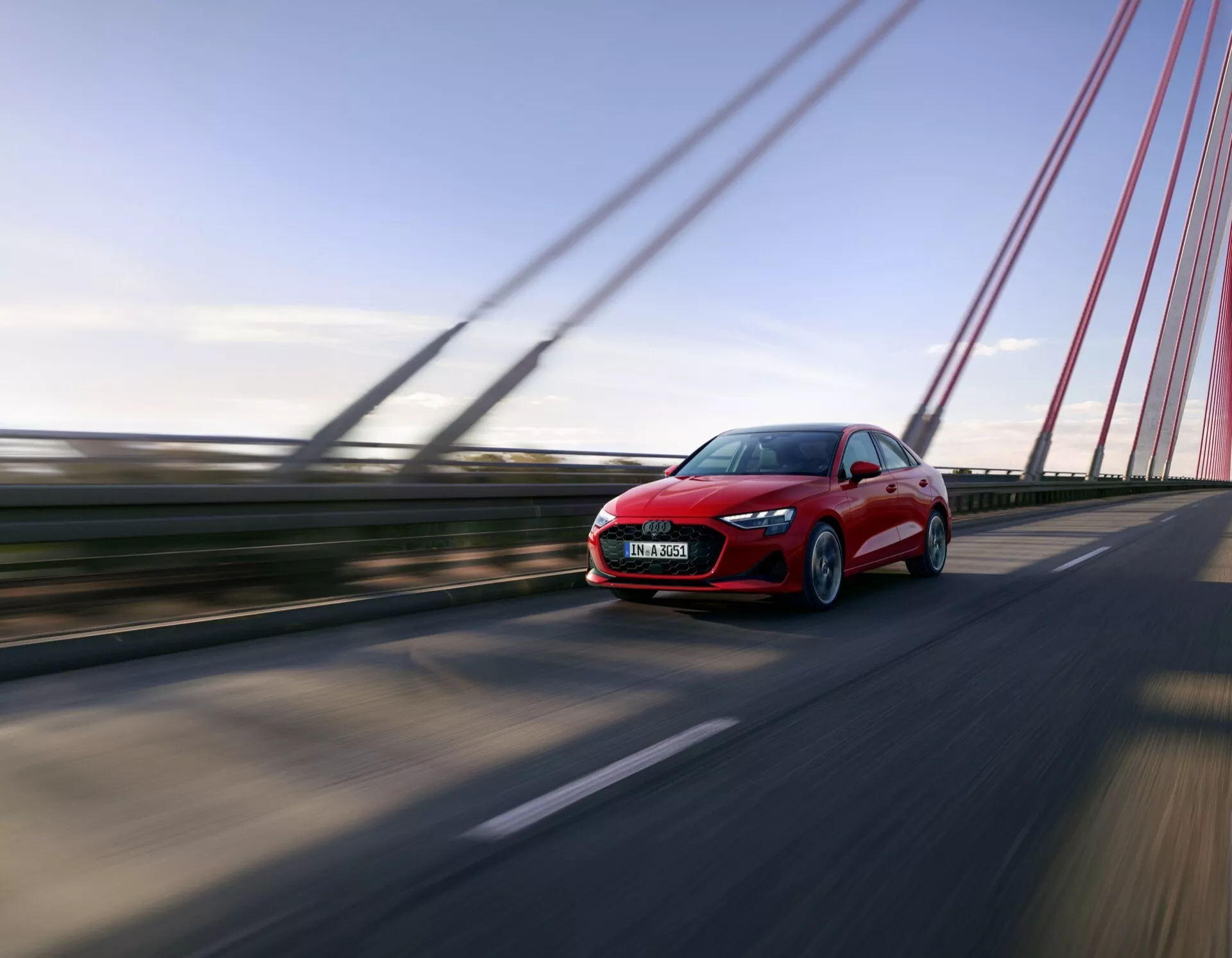Audi A3 (8Y-Facelift) 2024 v gibanju na mostu, ki prikazuje dinamičen pogled s sprednje in ene strani v nizkem kotu za dramatičen učinek, poudarjen z zameglitvijo gibanja okoli vozila, ki se hitro premika, z vidnimi oblikovalskimi elementi kot so LED žarometi in aerodinamične konture proti ozadju jasnega neba.