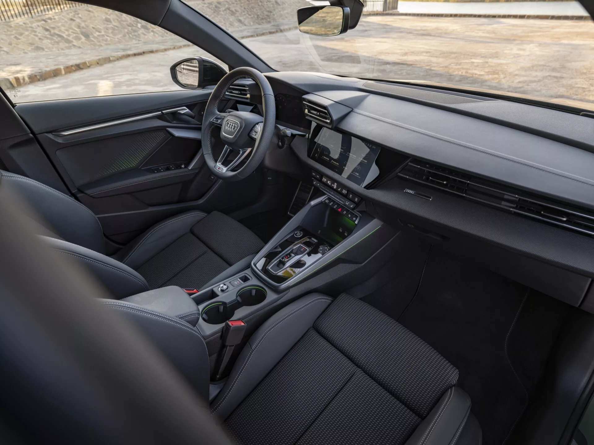 Pogled na notranjost Audi A3 (8Y-Facelift) iz leta 2024 iz perspektive zadnjega potnika, ki prikazuje elegantno armaturno ploščo z neprekinjenim digitalnim zaslonom, volan na levi strani z emblemon, minimalistične kontrole na sredinski konzoli, športno ergonomska prednja sedeža in napredne tehnološke funkcije
