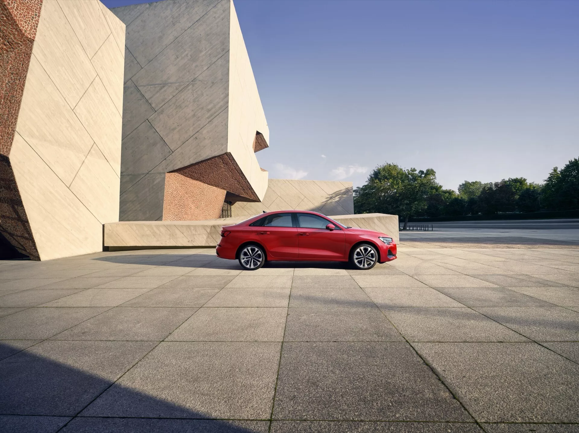 Facelift Audi A3 8Y 2024 v svetlo rdeči barvi, ki prikazuje avto iz sprednje bočne perspektive, izpostavljeno z elegantno zasnovo LED žarometov, sijočo sprednjo masko, aerodinamično oblikovanimi večkrakimi zlitinskimi kolesi in moderno silhueto na ozadju mestnega okolja z drznimi arhitekturnimi elementi, vse posneto v naravni sončni svetlobi