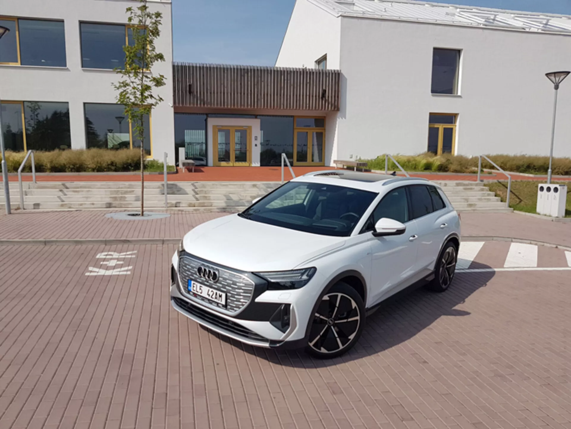 Beli Audi Q4 e-tron 2021, ki prikazuje svoje sodobne LED žaromete, šesterokotno sprednjo masko in velika lita kolesa, parkiran na tlakovanem območju z vzorcem opeke z urbanistično arhitekturo in polnilno postajo v ozadju, pod jasnim opoldanskim nebom.