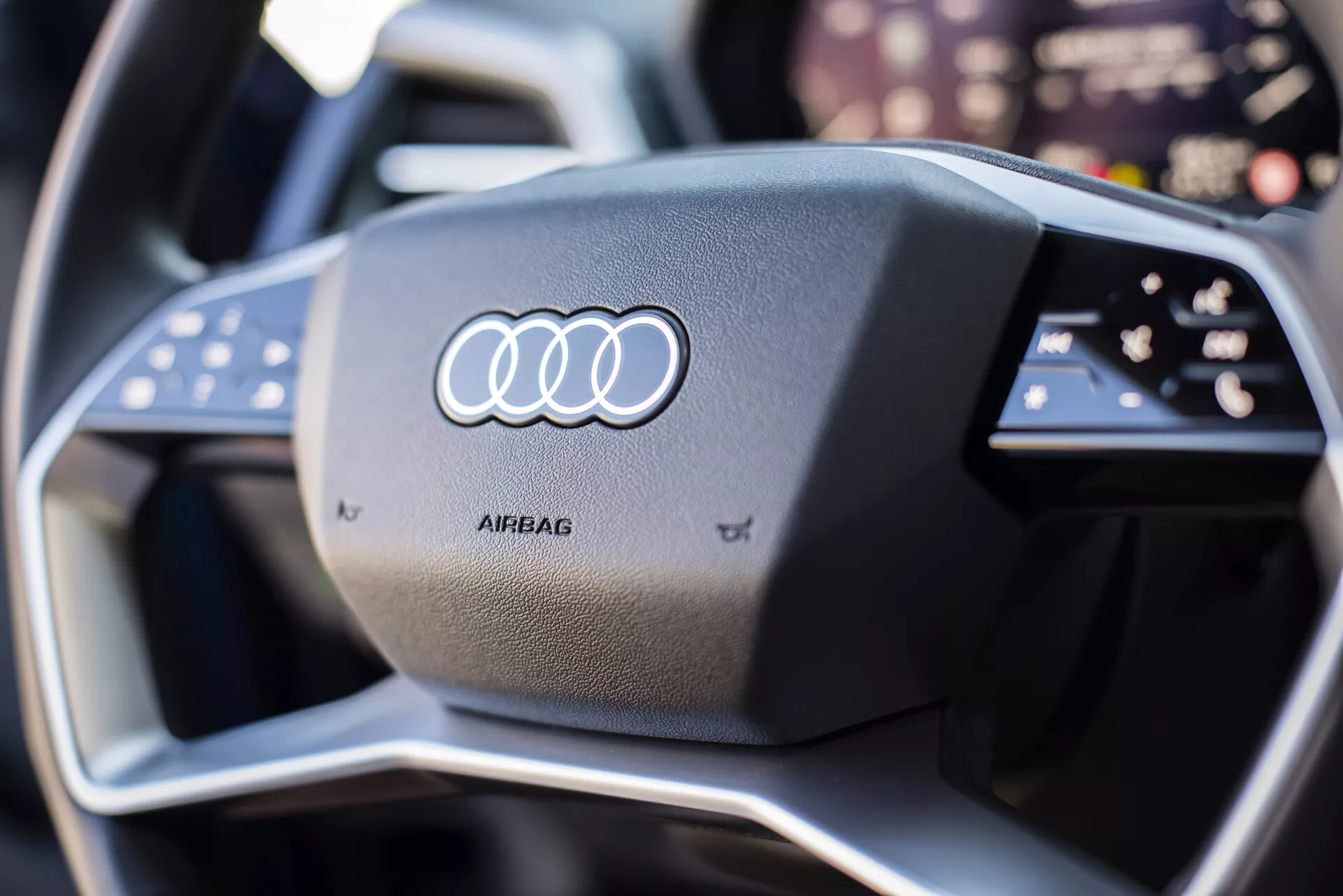 Posnetek iz bližine volana Audi Q4 e-tron 2021 z ikoničnim logotipom Audi na sredini za zračni meh, obdan z multifunkcijskimi gumbi, z moderno, športno zasnovo, ki vključuje kontrastne šive in namig na napredno tehnologijo digitalne armaturne plošče v ozadju