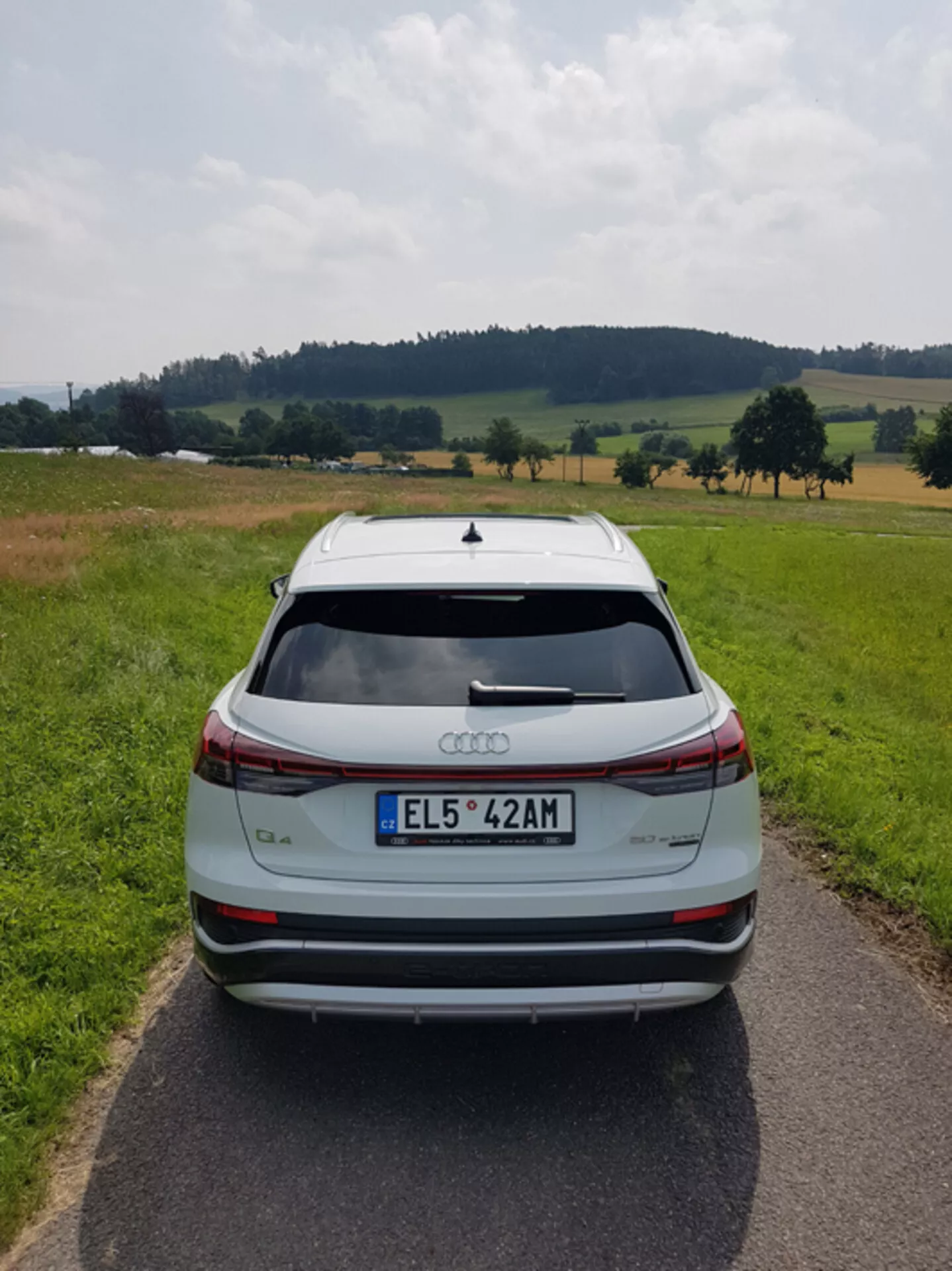 Zadnji pogled na srebrni Audi Q4 e-tron 2021 na ozki tlakovani cesti sredi pastoralne krajine z polji in drevesi pod delno oblačnim nebom, poudarjajoč njegov eleganten dizajn, zavorno luč v obliki letvice, zadek spojler, senzorje za parkiranje zadaj in dvobarvna lita platišča.