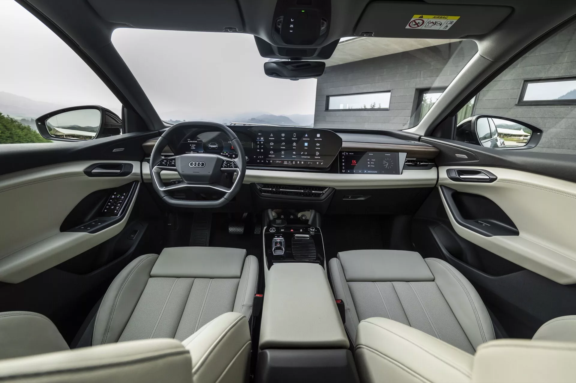 Notranji pogled na Audi Q6 e-tron 2024 iz perspektive zadnjega sedeža, s poudarkom na prednji vrsti. Svetlo usnjeni sedeži imajo nastavljive vzglavnike in elektronske nastavitvene kontrole. Instrumentna plošča prikazuje širok digitalni zaslon in velik osrednji zaslon na dotik za informacijsko zabavni sistem, obdan z gumbi za upravljanje klimatske naprave in načinov vožnje. Sredinska konzola vključuje držala za pijačo, shranjevalni prostor in prestavno ročico za avtomatski menjalnik. Volan je opremljen z več kontrolami ter panoramska streha nakazuje na obilico naravne svetlobe. Skupaj slika poudarja napredne tehnologije in luksuzne avtomobilske lastnosti.