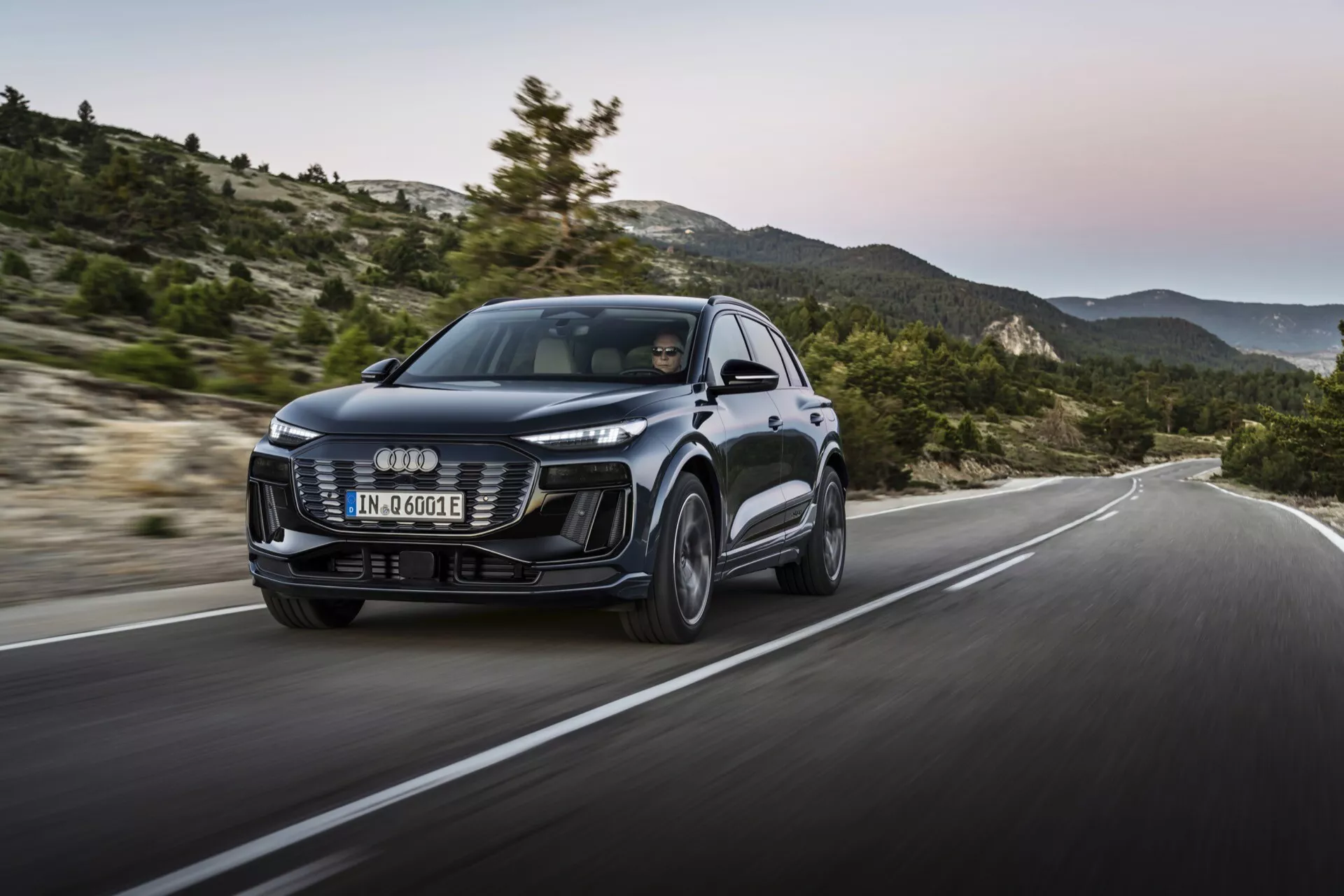 Audi Q6 e-tron 2024 z modernim dizajnom, veliko sprednjo masko, elegantnimi LED žarometi in izklesanimi linijami karoserije. Prednji tričetrtinski pogled z dinamičnim posnetkom v gibanju na hriboviti cesti, kar poudarja zmogljivost in vodljivost.
