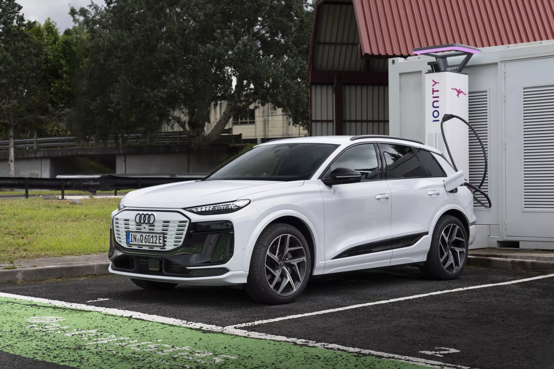 Pogled na SUV Audi Q6 e-tron 2024 bele barve iz tričetrtinske sprednje strani, ki poudarja sprednjo masko, LED žaromete, aerodinamične linije, večkraka kolesa in bočni profil. Vozilo je parkirano na označenem mestu za polnjenje električnih vozil, z vidnim polnilnim priključkom in polnilnikom IONITY v ozadju. Slika kontrastira dizajn avtomobila s temnim, oblačnim nebom in mokrim pločnikom ter prikazuje sodobno tehnologijo električnih vozil in napredno osvetlitev.