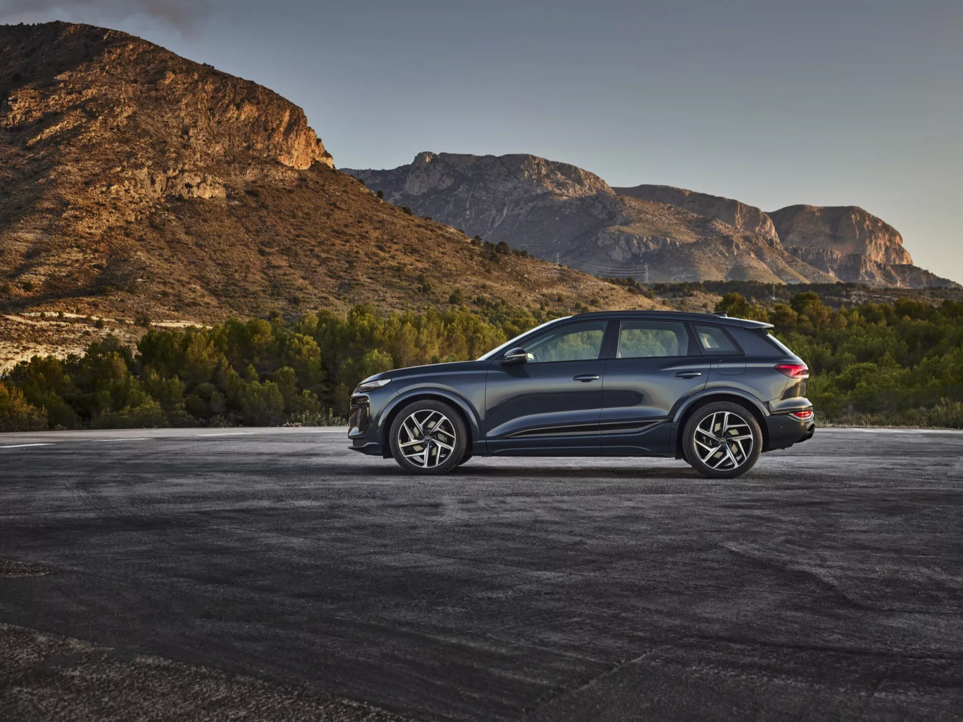 Moderen SUV, domnevno Audi Q6 e-tron 2024, parkiran na prostem s hribovitim ozadjem, zajet v tričetrtinskem zadnjem pogledu iz dvignjenega kota. Temna kovinska barva karoserije kontrastira z naravnim okoljem, poudarja elegantne linije in aerodinamične konture. Vidne podrobnosti vključujejo LED zadnje luči, velike aluminijaste platišča z večkrakovito zasnovo, dvojni izpušni sistem, zatemnjena okna, panoramsko strešno okno in zaščitne obloge vzdolž spodnjih robov. Zadnje okno je rahlo nagnjeno s zadnjim spojlerjem in velikimi ogledali, ki imajo verjetno vgrajene smerokaze, kar povečuje športni in luksuzni videz vozila.