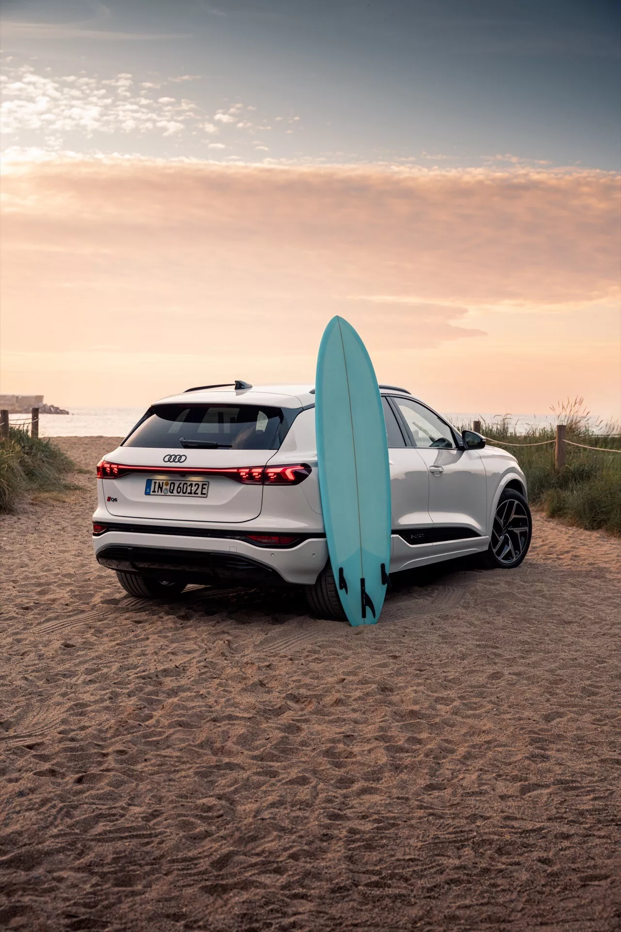 Slika prikazuje bel Audi Q6 e-tron 2024 SUV, parkiran na peščenem terenu ob plaži s surfarsko desko. Posneta je iz zadnjega trikotnega kota, ujame elegantne linije avtomobila, LED zadnje luči, parkirne senzorje, vzvratno kamero ter večkraka lita platišča. Odsev sončnega zahoda poudarja napredno kakovost barve, medtem ko deska za deskanje naslonjena na avtomobil prikazuje element pustolovskega življenjskega stila.