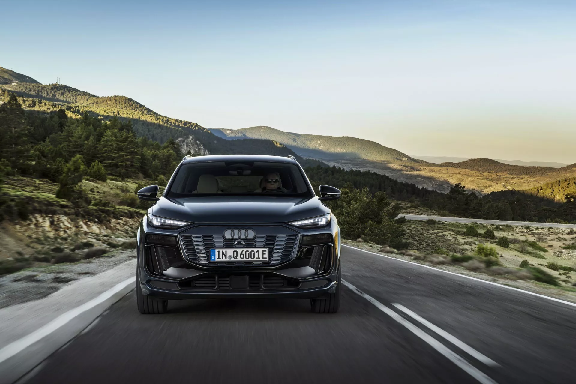 Frontalni pogled na Audi Q6 e-tron 2024, ki prikazuje njegovo sprednjo mrežo, žaromete in odbijač s štirimi prepletenimi obročki kot logotipom. Evropska registrska tablica je nameščena na mreži, in oznaka 'e-tron' označuje njeno električno/hibridno naravo. Avto se vozi po odprti cesti z gorsko pokrajino v ozadju, kar nakazuje nastavitev poznega popoldneva. Lastnosti vključujejo elegantne LED žaromete, estetsko mrežo, senzorje/kamere za pomoč vozniku, aerodinamična ogledala in učinkovit dizajn.