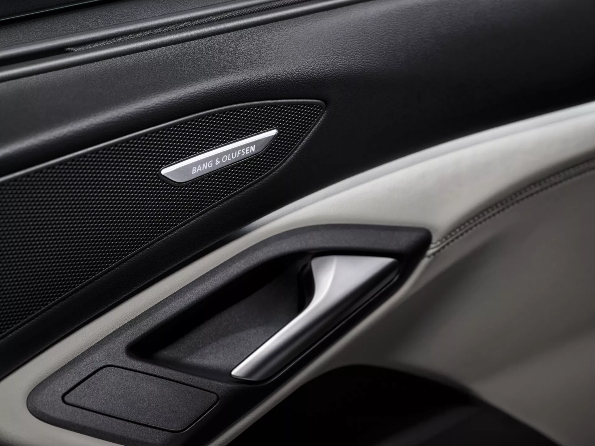 Podroben posnetek notranje plošče vrat Audi Q6 e-tron 2024 GF, ki poudarja vrhunske lastnosti, kot so rešetka zvočnika Bang & Olufsen, kromiran ročaj vrat in kombinacija usnjenih in karbonskih tekstur. Dobro osvetljena slika se osredotoča na podrobnosti plošče, prikazuje luksuzne in tehnološke elemente vozila.