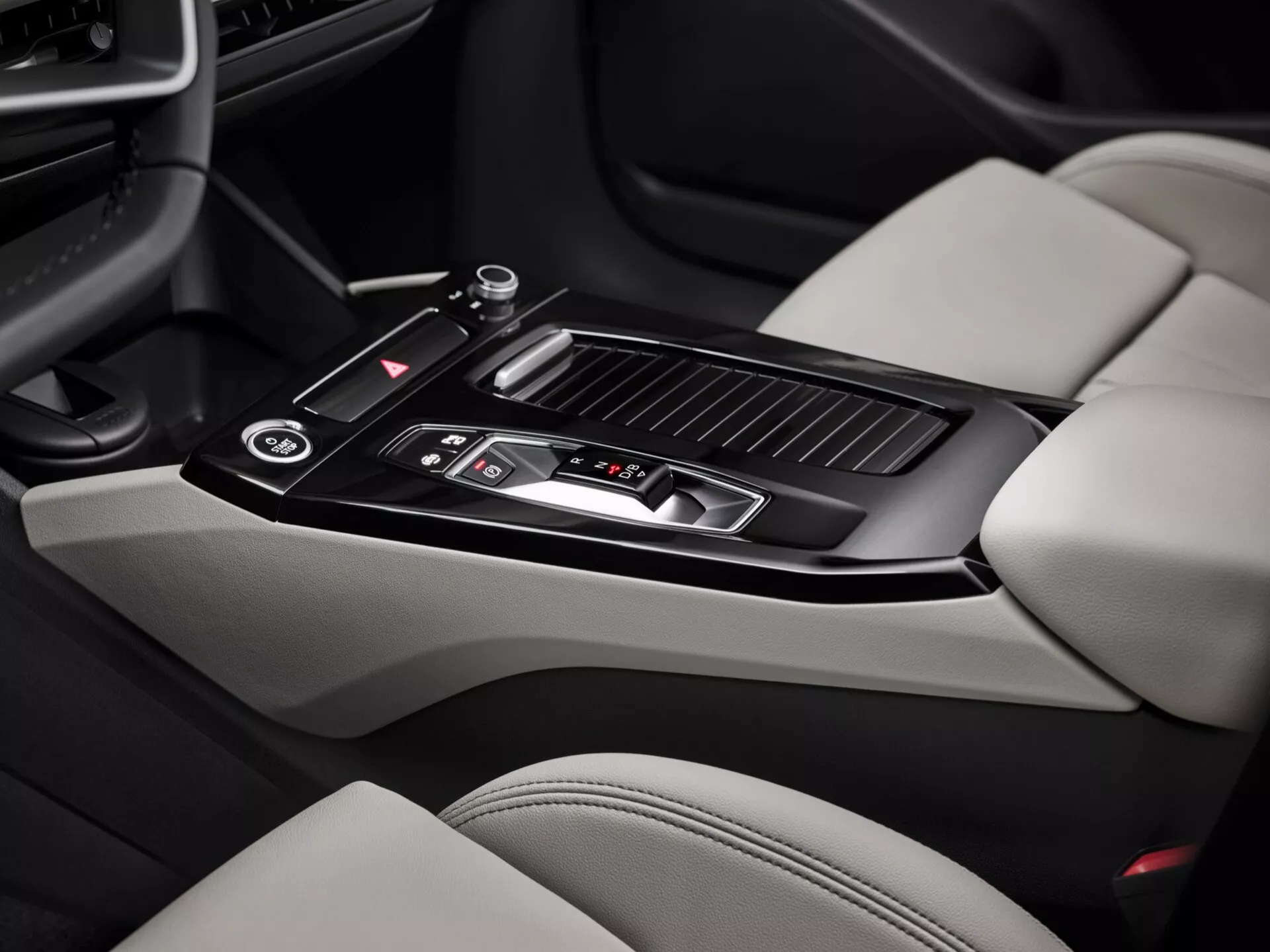 Notranji posnetek Audi Q6 e-Tron 2024, osredotočen na območje osrednje konzole. Sliko, posneto iz potnikove perspektive, odlikuje sijajen črn prestavni ročaj z kovinskimi detajli, več nadzornih gumbov, majhno shranjevalno območje s pomičnim pokrovom, dva nosilca za skodelice, usnjen naslon za roke, zračniki za nadzor klime in deli svetlo obarvanih usnjenih sedežev s kontrastnimi temnejšimi deli. Značilnosti osrednje konzole prikazujejo sodoben dizajn in luksuzne elemente.