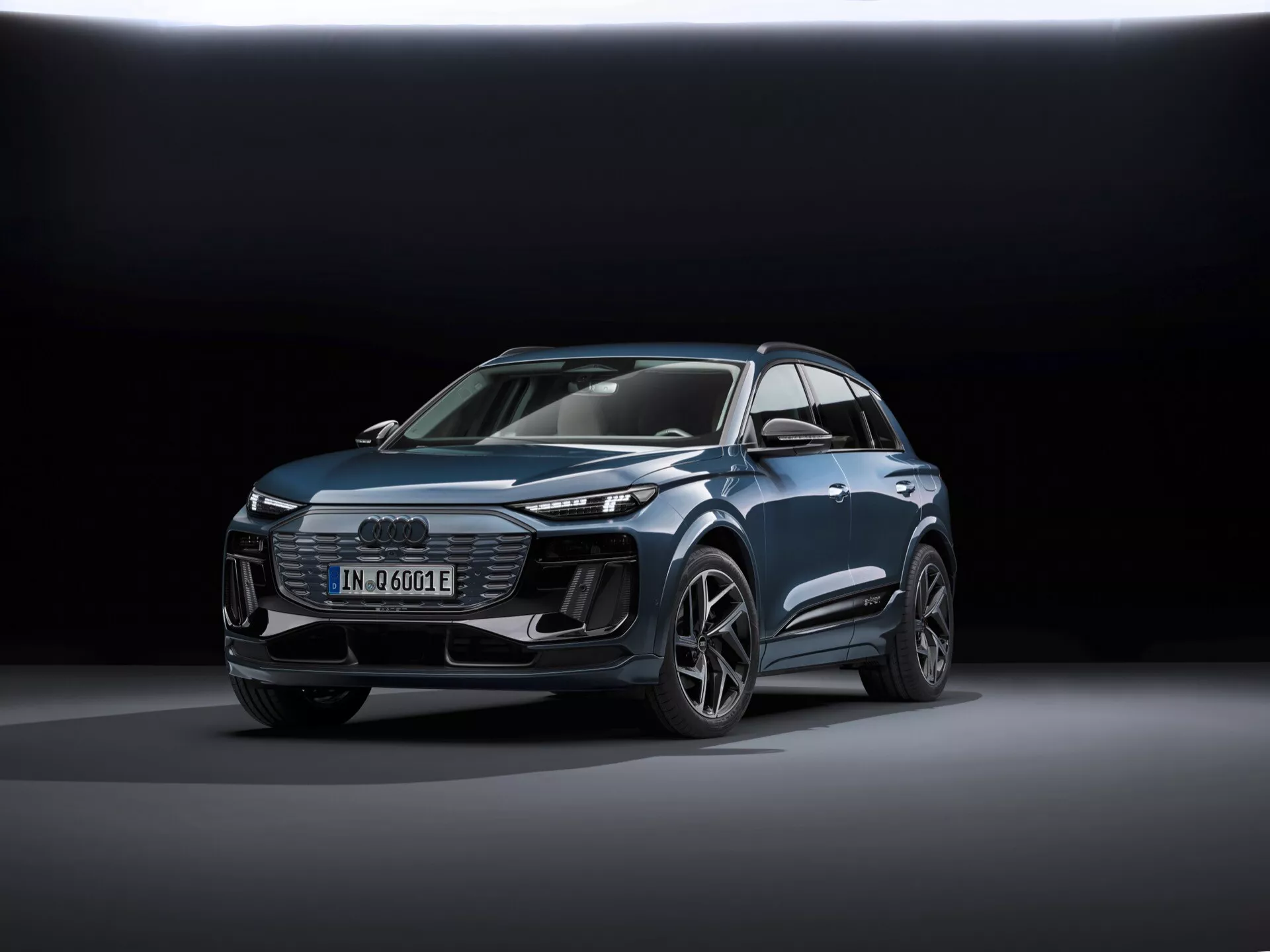 Slika prikazuje sodoben SUV Audi Q6-e-tron 2024 iz treh četrtin spredaj, s poudarkom na sprednjem in stranskem profilu. Glavni identifikatorji vključujejo značko štirih obročev Audi na mreži in značko 'e-tron', ki označuje električni model. Posnetek je narejen iz nizke perspektive, kar poudarja držo vozila in prikazuje njegove oblikovne linije, mišičaste blatnike in aerodinamične lastnosti. Avto je postavljen v središče okvirja, stoji na gladki odsevni površini z mehko, razpršeno osvetlitvijo in temnim gradientnim ozadjem, kar poudarja oblikovanje vozila. Vidne tehnološke lastnosti vključujejo LED žaromete, dnevne luči (DRL), digitalni zaslon in zlitinska kolesa s kompleksno zasnovo. Celotna sijoča predstavitev nakazuje promocijsko fotografijo, ki poudarja tehnologijo, dizajn in luksuzne vidike vozila.