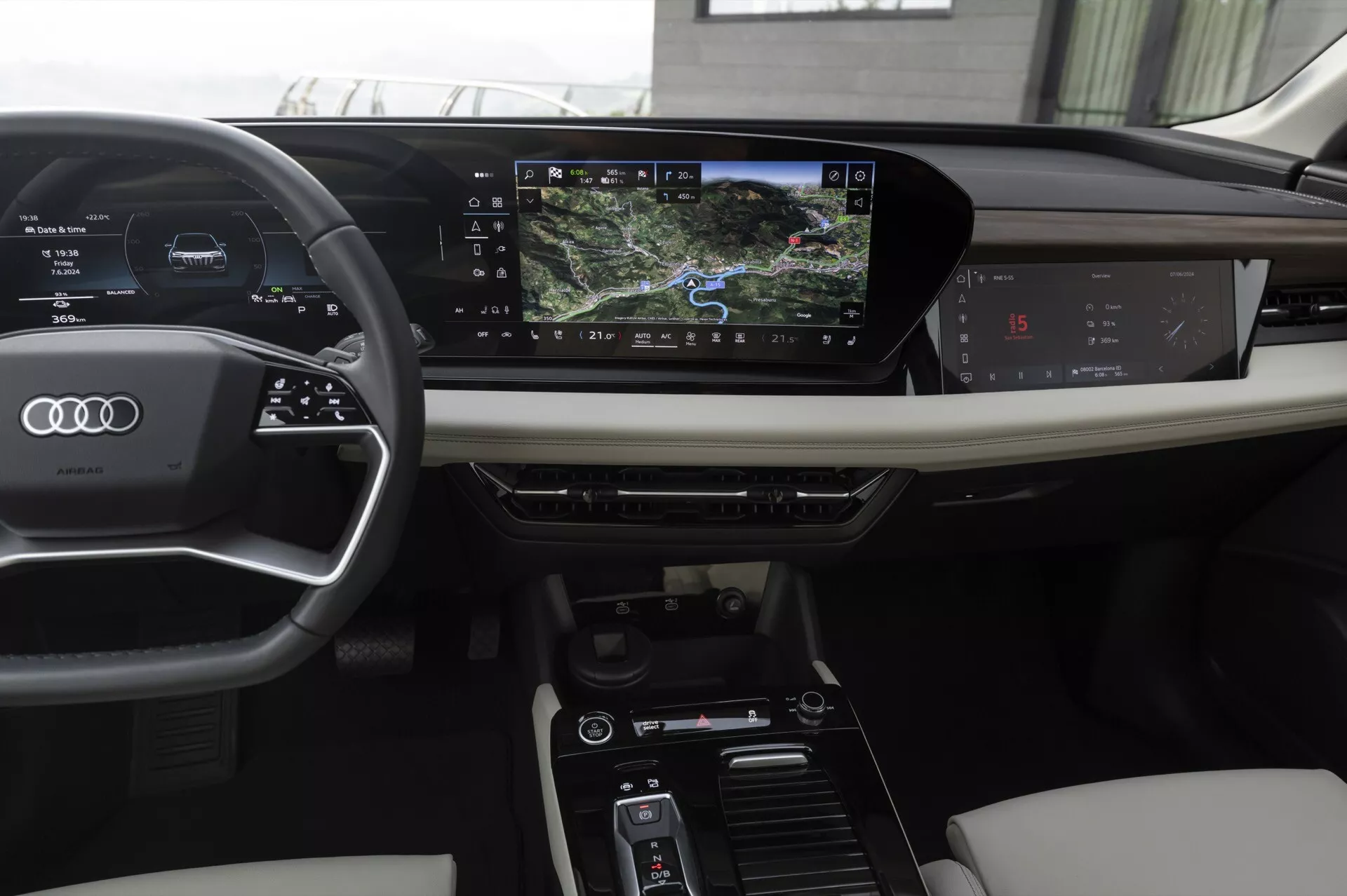 Notranjost sodobnega Audi Q6 e-tron 2024, prikazana z voznikove perspektive. Značilnosti vključujejo volan s tremi kraki, oblečen v usnje, z nameščenimi kontrolami, digitalno instrumentno ploščo, ki prikazuje hitrost in navigacijo, ter horizontalno usmerjen zaslon na dotik infotainment sistema. Osrednja konzola vključuje klimatske nastavitve in načine vožnje. Luksuzni materiali, kot sta usnje in leseni okras, izboljšujejo estetiko.