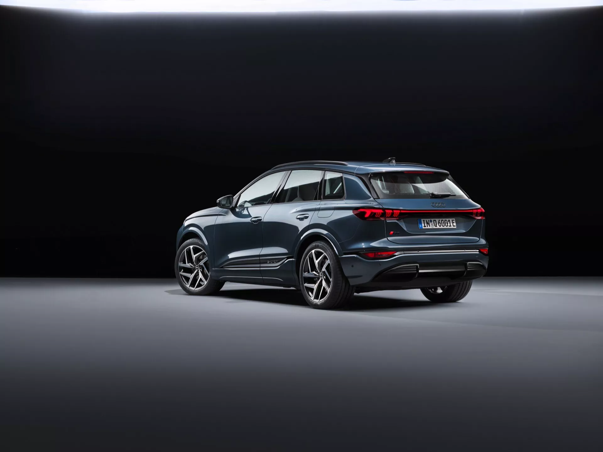 Zadnji trikotni pogled na Audi Q6 e-tron 2024 (gf) prikazuje sodoben SUV, postavljen diagonalno na gladki, odsevni talni površini v salonu. Ozadje prehaja iz temnega v svetlo, kar poudarja vozilo. Vidne lastnosti vključujejo povezano zadnjo svetlobno skupino, značilni spojler na strehi, izrazite koloteke z velikimi platišči iz lahkih kovin, kromirano obrobo vzdolž oken, zadnji odbijač z odsevniki in senzorji ter dvojni izpušni sistem, ki nakazuje na zmogljivost, kar lahko pomeni, da je model hibriden ali električen.