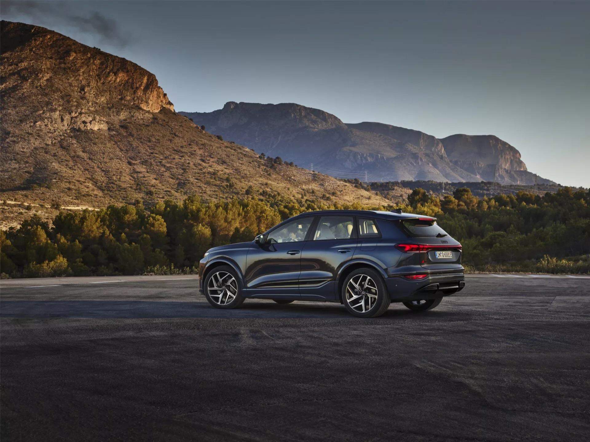 Slika prikazuje sodoben SUV Audi Q6 e-Tron 2024 v zunanjem okolju s slikovitim gorskim ozadjem med zoro ali mrakom. Treh četrtinski zadnji kot poudarja značilne LED zadnje luči, čist dizajn zadnjih vrat, ki verjetno vključuje zadnjo kamero, in dvojni izpušni sistem. Stranski profil prikazuje večkrake aluminijaste platišča, velike okenske površine in nagnjeno strešno linijo, kar nakazuje športno ali luksuzno opremo z možnimi aerodinamičnimi lastnostmi. Stranska ogledala bi lahko vključevala tehnologijo nadzora mrtvih kotov. Naravna osvetlitev povečuje privlačnost vozila, kar kaže na povezavo z avanturo in zmogljivostjo.