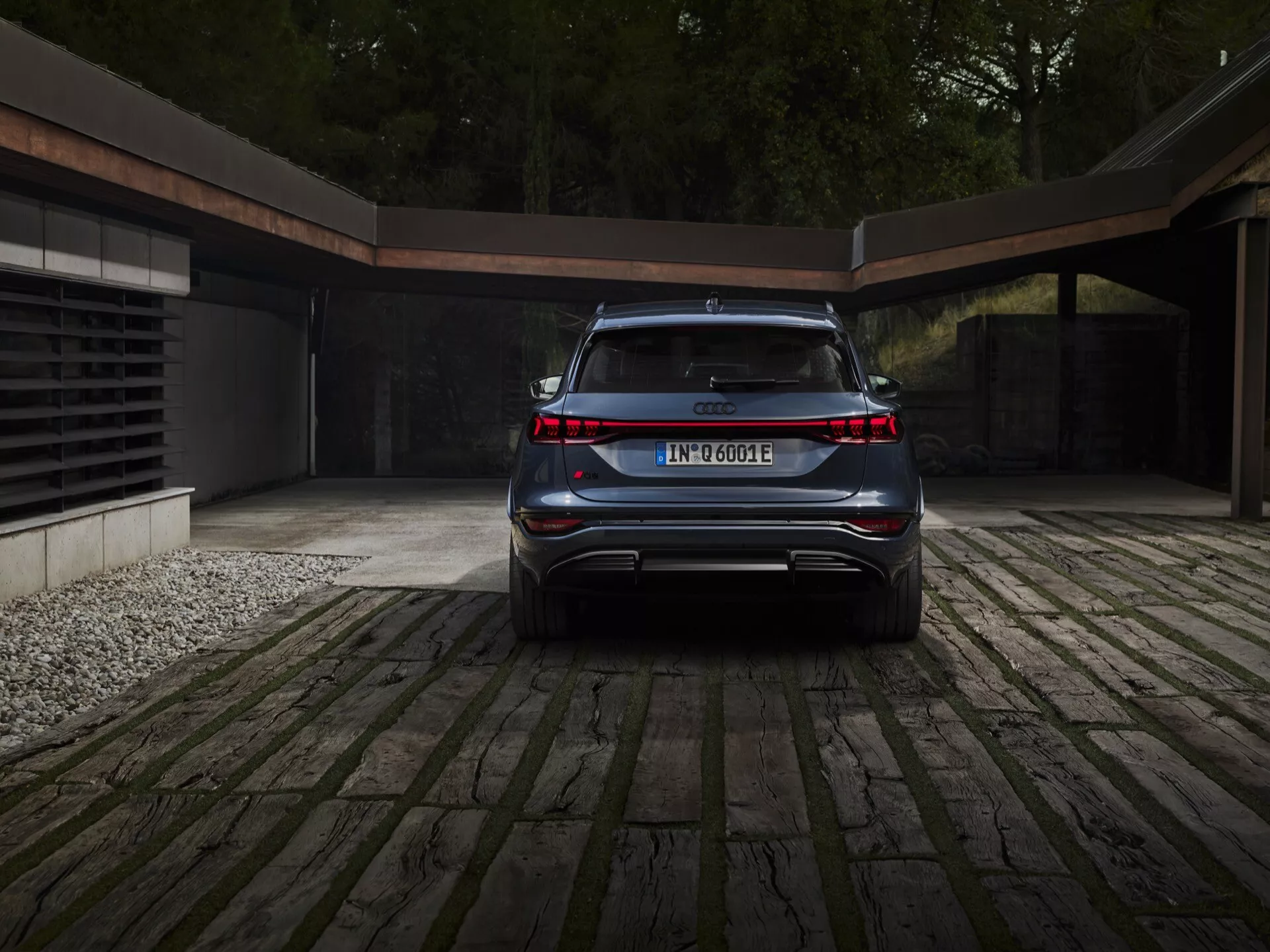 Zadnji kotni pogled na Audi Q6 e-tron 2024, ki poudarja njegovo čisto, elegantno in moderno zasnovo. Vidne ključne lastnosti vključujejo neprekinjen svetlobni trak, ki povezuje obe zadnji luči, oznako modela in električnega pogonskega sistema, integriran zadnji odbijač s športnim difuzorjem, naprej nagnjeno zadnje steklo, ki nakazuje SUV ali crossover, skrite izpušne cevi in osrednje nameščeno registrsko tablico. Avto je parkiran na tlakovani dovozni poti z namigom sodobne garaže v ozadju, kar nakazuje luksuzen in okoljsko osveščen življenjski slog.