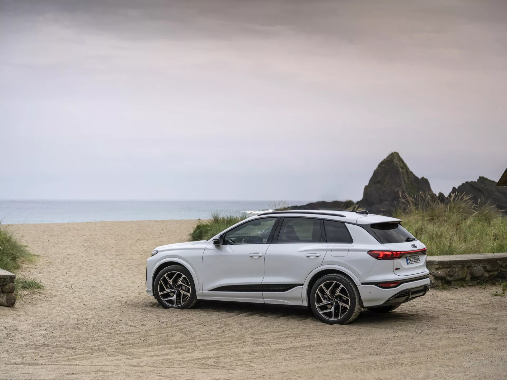 Beli SUV Audi Q6 e-tron 2024 je parkiran na makadamski cesti z ozadjem podobnim plaži, ujet iz zadnjega trikotnega pogleda. Slika poudarja sodobne oblikovalske elemente vozila, vključno z zadnjimi lučmi, zadnjim odbijačem, elegantnimi kolesi in elegantnimi linijami. Večja oddaljenost od tal in splošen drža nakazujeta primernost za različne terene. Mehka, enakomerna osvetlitev kaže na oblačen dan ali obdobje blizu sončnega vzhoda/zahoda, kar poudarja pustolovsko okolje. Vidna so sodobna litjeva kolesa, zadnji spojler in LED zadnje luči, kar nakazuje napredno tehnologijo in športni značaj.