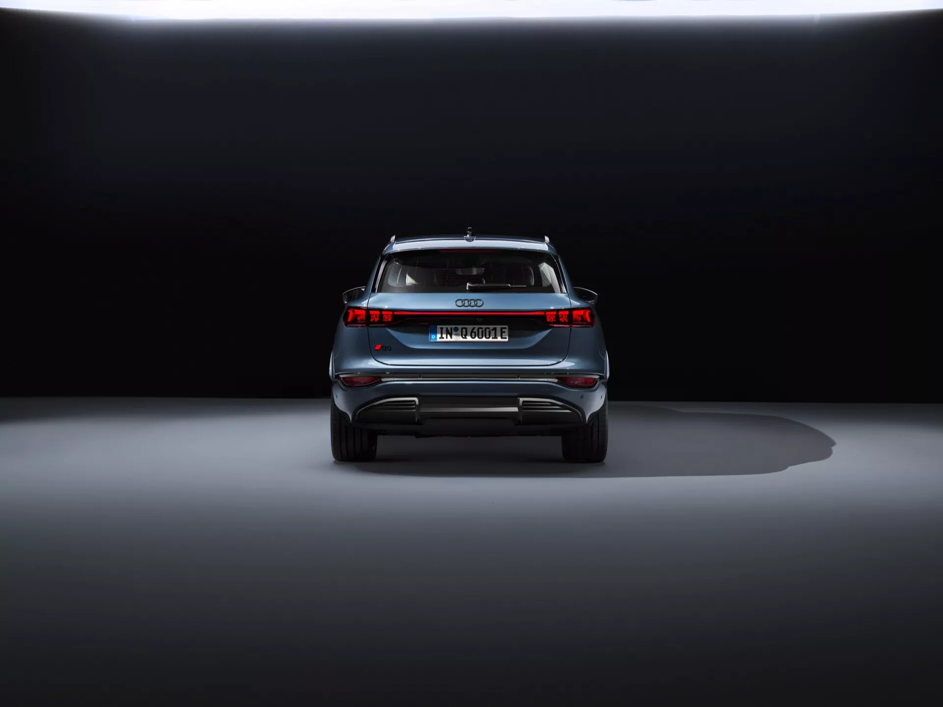 Pogled od zadaj na Audi Q6 e-tron 2024 na razstavi s dramatično osvetlitvijo. Posnetek iz talne ravni poudarja sodobne LED zadnje luči, pomanjkanje izpušnih cevi, široko svetlobno vrstico, evropsko registrsko tablico, strešni spojler in odsevne varnostne elemente, kar kaže na luksuzno električno ali hibridno vozilo