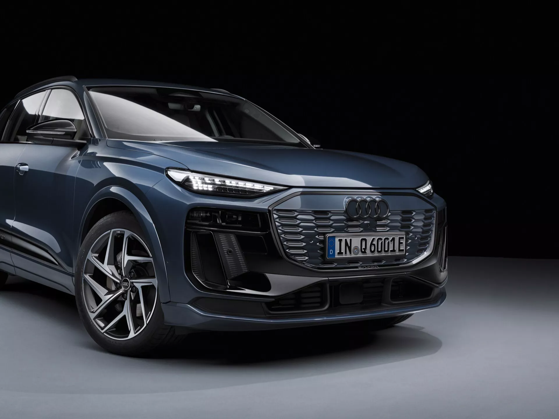 Slika prikazuje sodobno Audi Q6 e-tron 2024 v studijskem okolju, prikazano s prednjega tričetrtinskega pogleda, kar daje jasen vtis o oblikovanju vozila spredaj in s strani. Jasno je videti levi sprednji kot avtomobila, vključno s sprednjo masko s satastim vzorcem in osrednje nameščenim znakom Audi, kompleksnimi LED žarometi, dodatnimi dovodi zraka ali senzorji pod žarometi, športnim sprednjim odbijačem, velikim aluminijastim kolesom z nizkoprofilno pnevmatiko in orisom bočnega ogledala. Slika poudarja oznako elektrificiranega modela ('e') ter izpostavlja njegove oblikovne in tehnološke značilnosti. Ozadje je preprosto in temno, kar zagotavlja, da ni motenj od samega vozila.