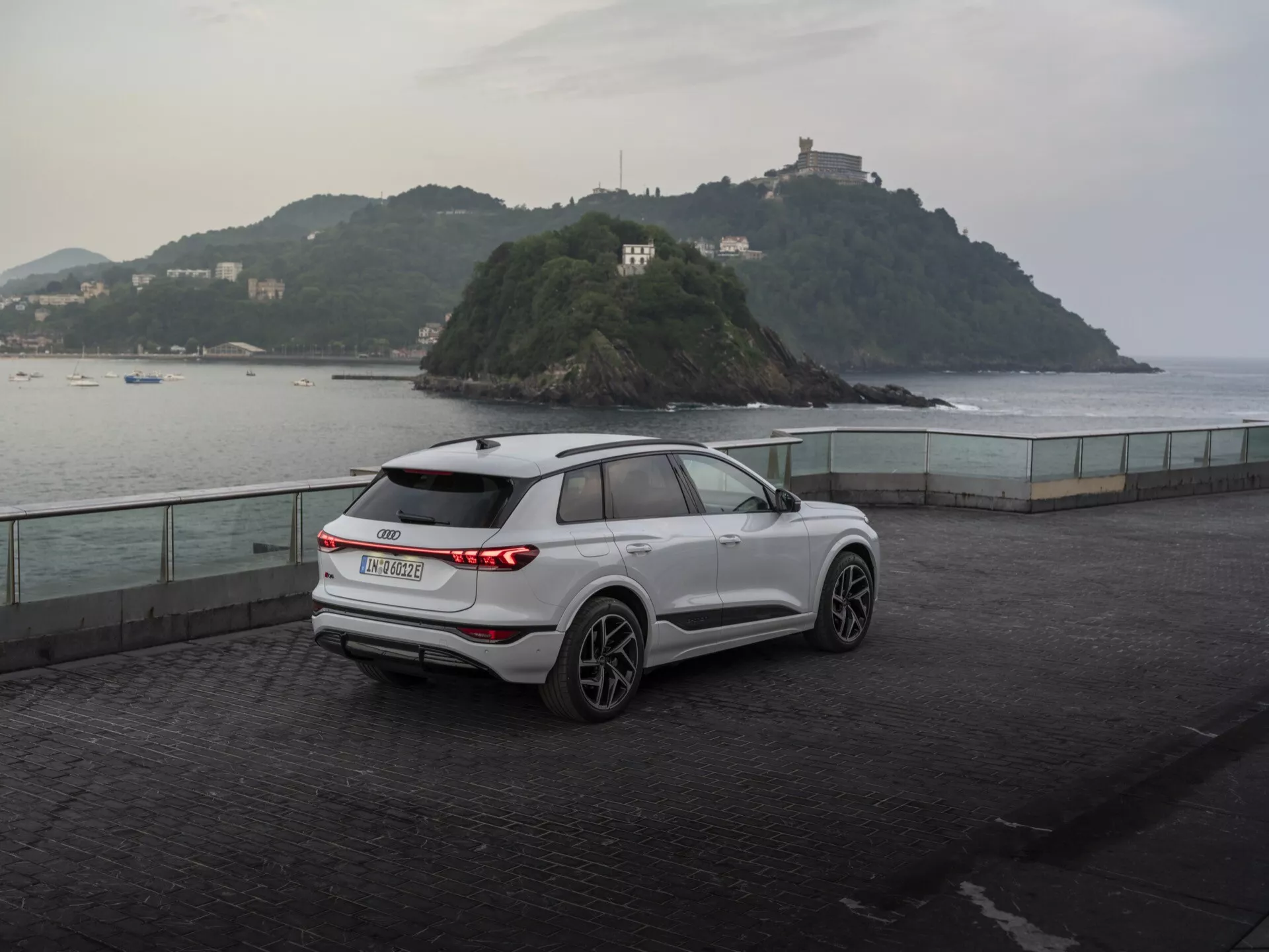 Tričetrtinski zadnji pogled na moderni, beli SUV Audi Q6 e-tron 2024, parkiran na obalni promenadi ob mraku, ki prikazuje horizontalne zadnje LED luči, proizvajalčev znak, zadnji brisalec, petkraka aluminijasta platišča, ogledala z vgrajenimi smerniki, strešne nosilce in oblikovane linije karoserije proti mirnemu ozadju z majhnim otokom in stavbo, kar poudarja mešanico uporabnosti in zmogljivosti.