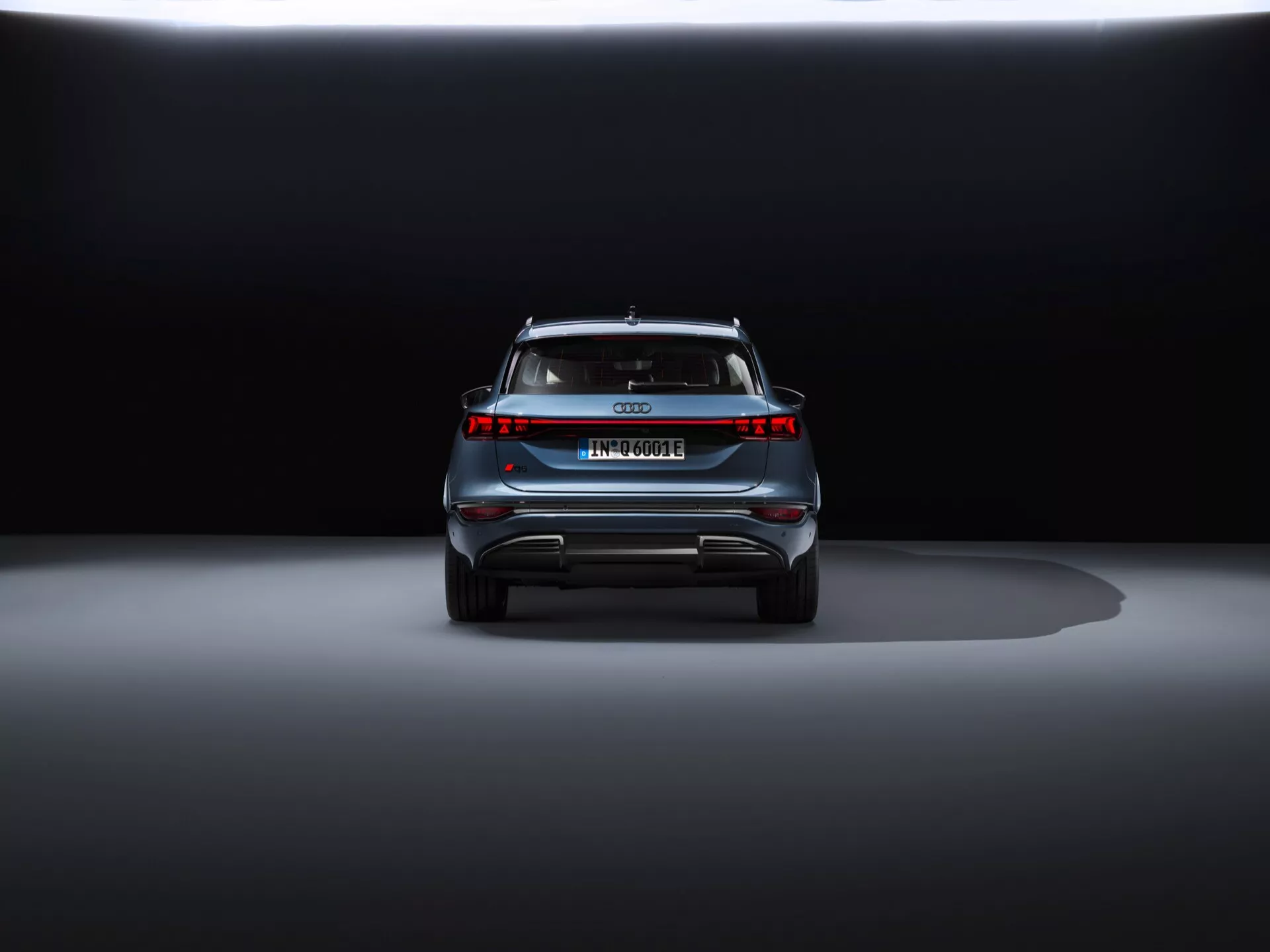 Zadnji pogled na Audi Q6 e-tron 2024 proti temnemu ozadju, ki prikazuje eleganten dizajn, osrednji emblem, ime modela, vodoravno svetlobno vrstico, zadnji spojler in odbijač v slogu difuzorja z odsevnimi elementi.