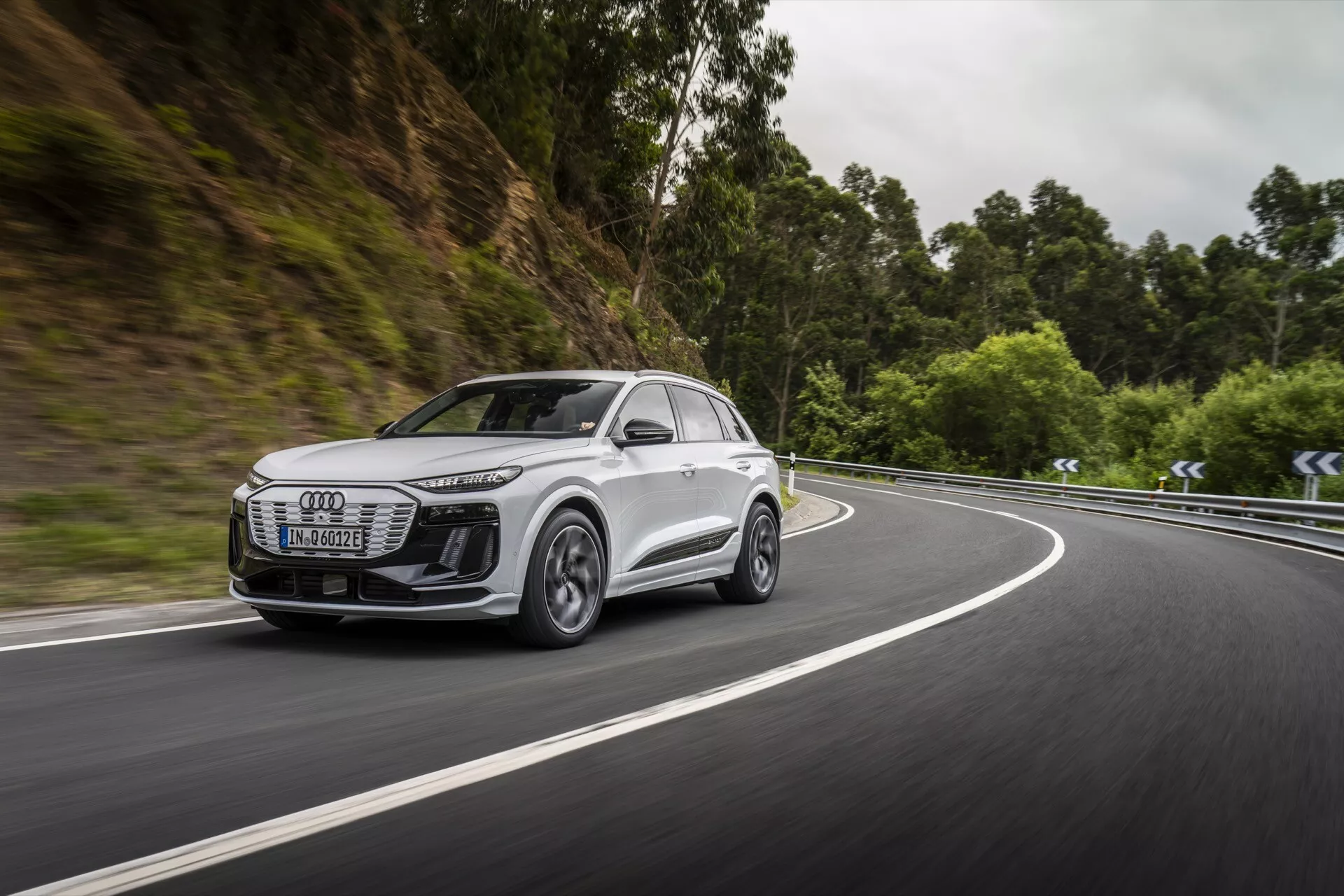 Slika zajame dinamičen stranski sprednji pogled na sodoben bel SUV, verjetno model Audi Q6 e-tron 2024, ki potuje po cesti obkroženi z gozdnatim območjem. Poudarek je na sprednji rešetki, žarometih, bočnem profilu, kolesih in aerodinamiki stranskih ogledal vozila. Žarometi so elegantni s tehnologijo LED, kar nakazuje napredno osvetlitev. Kolesa iz aluminijeve zlitine z dizajnom z več naperami nakazujejo zmogljivosti visokega razreda. Premikanje avtomobila na ukrivljeni cesti poudarja njegove sposobnosti upravljanja in možne napredne sisteme pomoči pri vožnji. Zamegljeno ozadje poudarja hitrost in učinkovitost pri obvladovanju zahtevnih situacij.