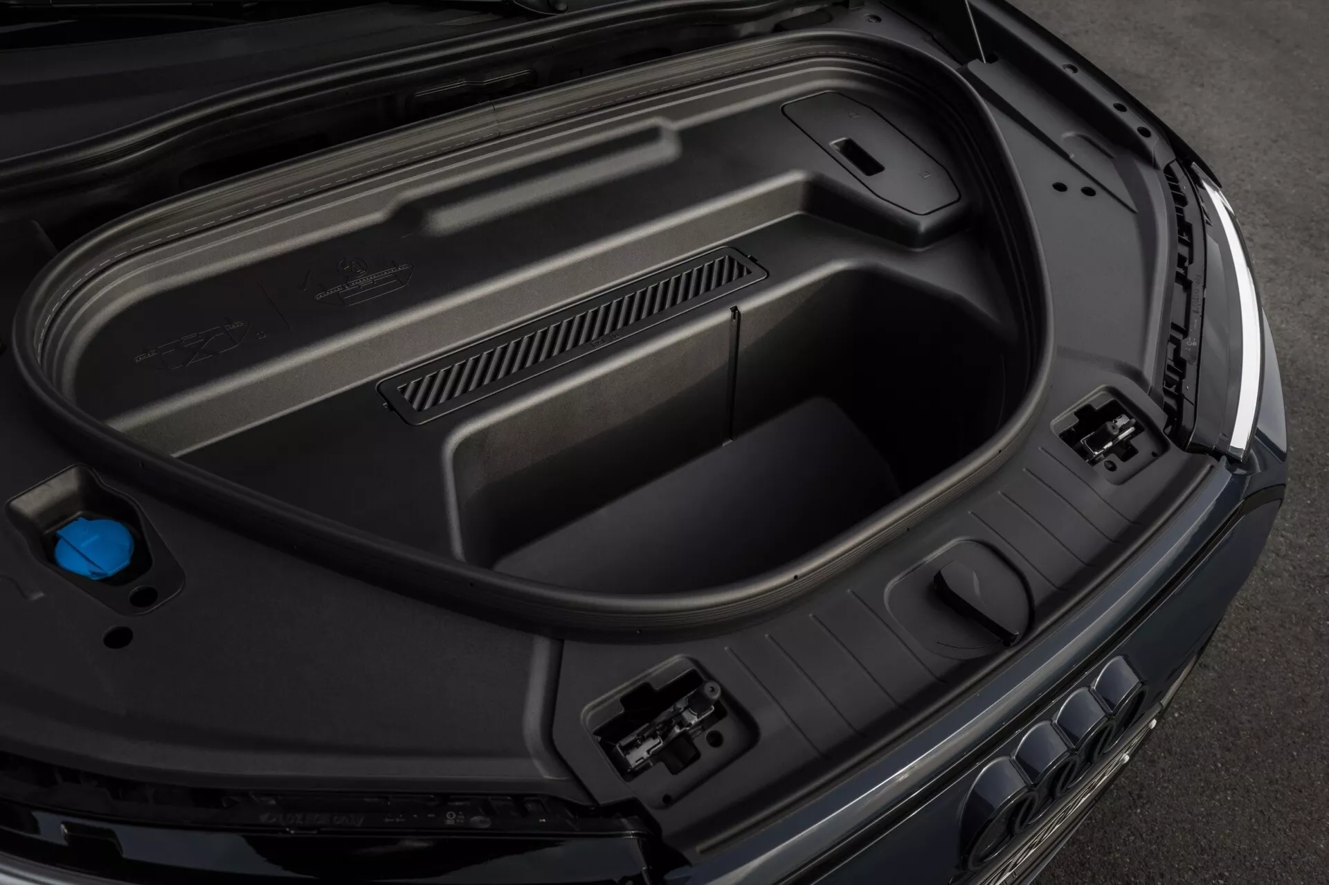 Posnetek od blizu iz visoke perspektive zadnjega prtljažnega prostora Audi Q6-e-Tron 2024, ki prikazuje notranje lastnosti, vključno z oblikovanimi plastičnimi ploščami za vzdržljivost, vtičnicami za dodatke na desni strani, vgrajenim shranjevalnim prostorom na dnu in modrim pokrovčkom na levi, kar kaže na napredno avtomobilsko tehnologijo in elegantno, premium končno obdelavo.