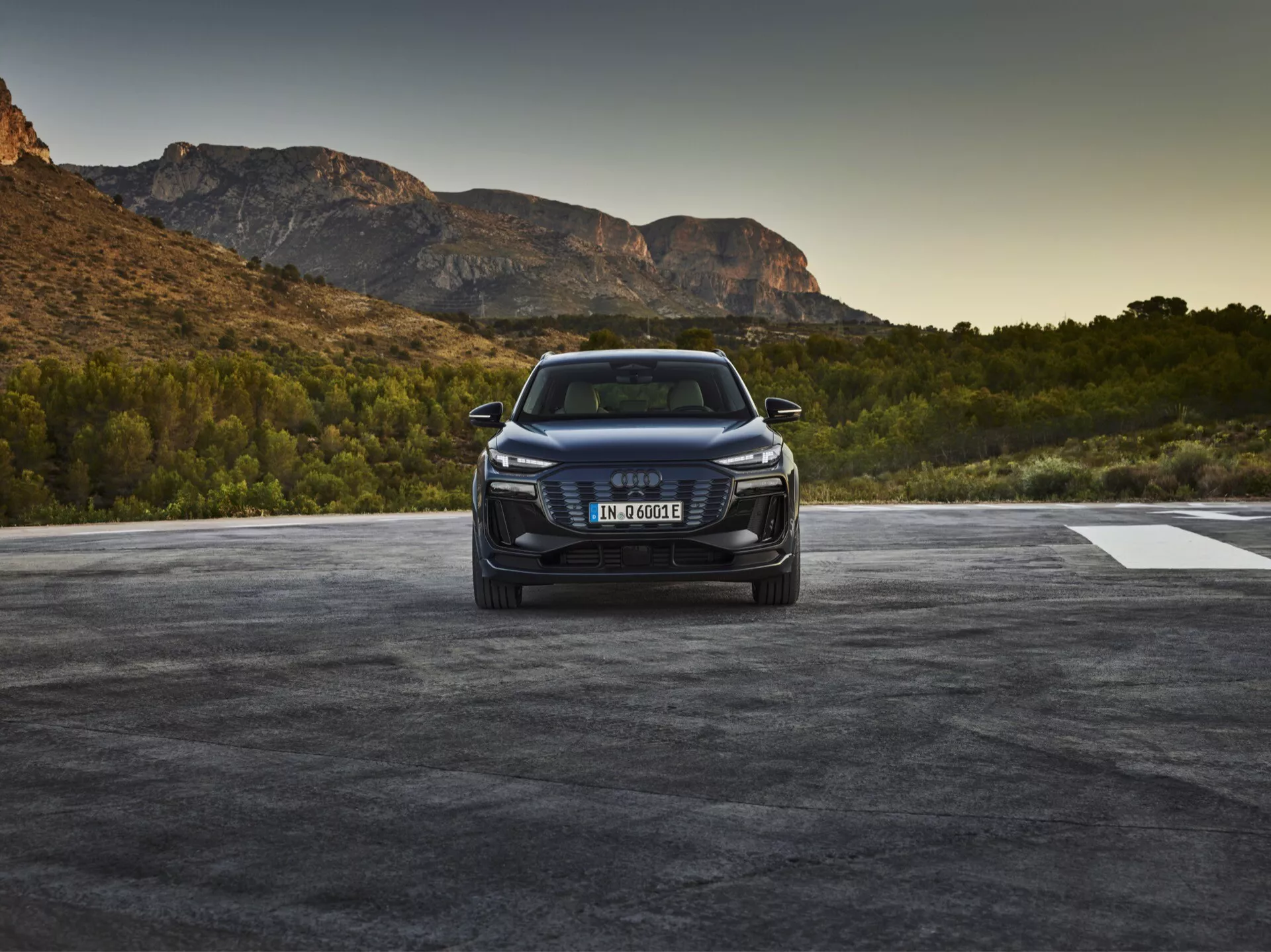 Slika Audija Q6 e-tron 2024, posneta iz sprednjega tričetrtinskega kota, prikazuje sprednjo masko vozila z emblemon znamke, ostre LED ali laserske žaromete ter podroben sprednji odbijač z zračnimi rešetkami in po možnosti senzorji ali kamerami, ki nakazujejo sposobnosti asistence vozniku. Pokrov motorja ima obrisne linije, eleganten stranski profil pa nakazuje aerodinamiko z dvignjeno držo SUV-ja. Vozilo je postavljeno v naravno okolje, ki namiguje na sončni vzhod ali zahod.