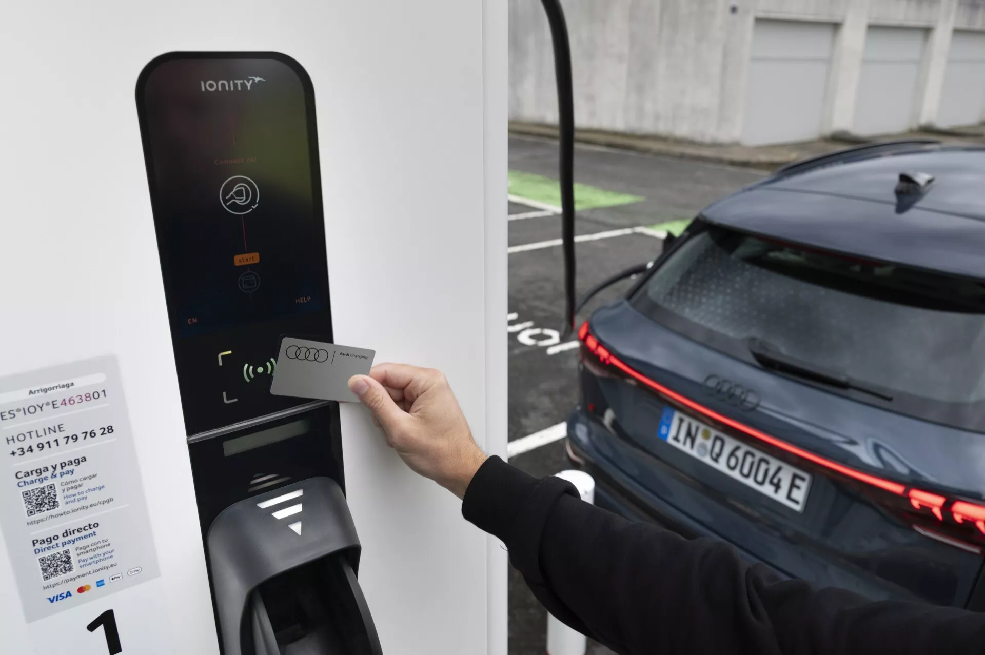 Slika osebe, ki drži RFID kartico blizu čitalnika polnilne postaje za električna vozila IONITY, usmerjena v zadnji del sodobnega temno obarvanega avtomobila s horizontalnimi LED zadnjimi lučmi, kar kaže na njegovo električno naravo. Avto, verjetno EV, je parkiran poleg polnilnika, kar kaže, da je v postopku polnjenja ali se pripravlja na polnjenje.