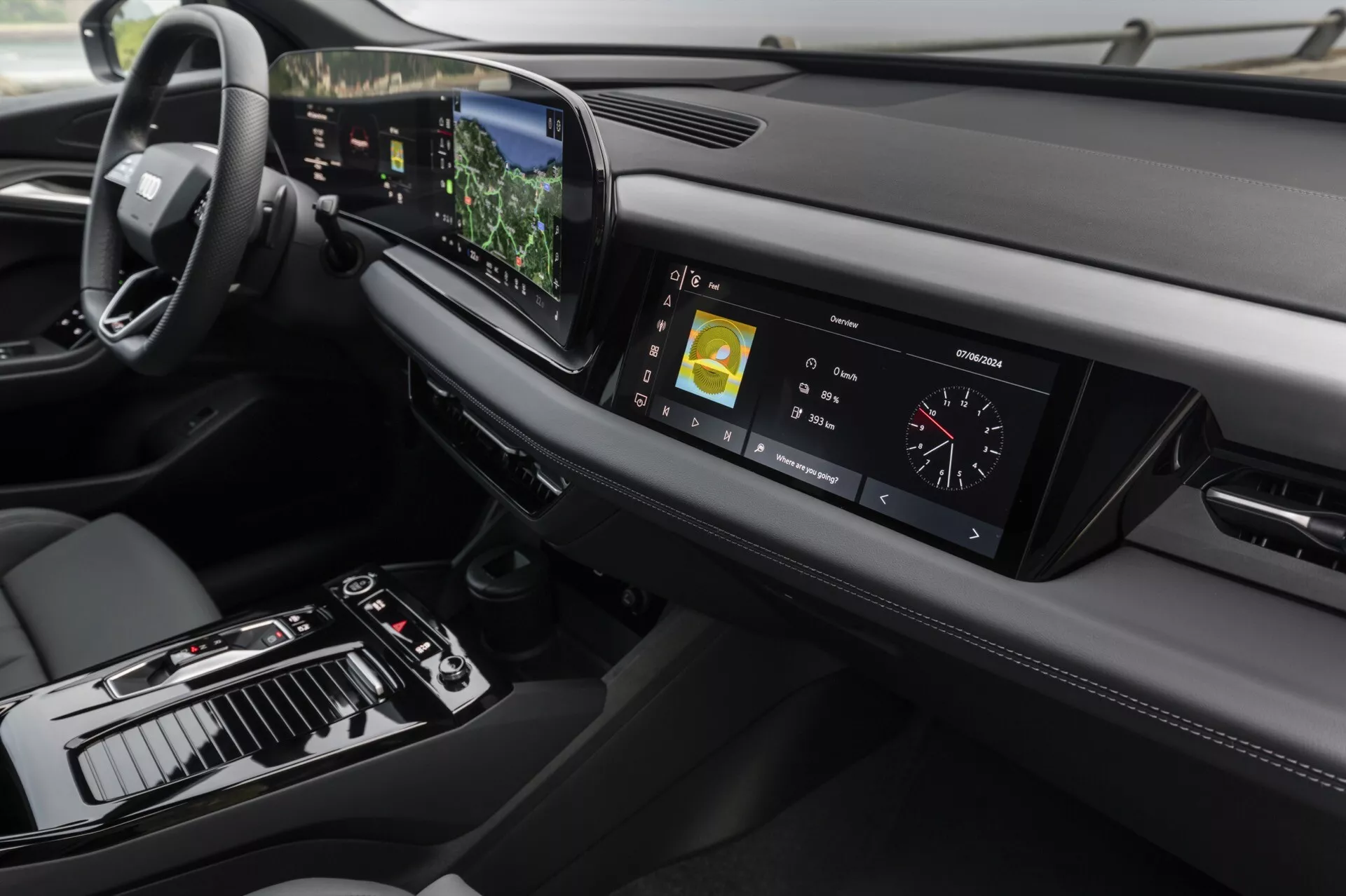 Pogled na notranjost Audija Q6 e-tron 2024, osredotočen na armaturno ploščo in osrednjo konzolo iz rahlo povišanega kota. Na levi je viden volan, kar kaže na konfiguracijo za vožnjo na levi strani, medtem ko osrednja konzola vsebuje prestavno ročico, različne kontrole in velik zaslon na dotik. Digitalni instrumentni zaslon prikazuje vozne meritve. Elegantna armaturna plošča se vodoravno razteza z zračniki in drugim oblikovnim elementom na desni strani. Napredna tehnologija je očitna na osrednjem zaslonu na dotik, ki prikazuje navigacijske in večpredstavnostne informacije, na projekcijskem zaslonu, vidnem nad volanom, in na tipkah na dotik za nadzor klimatske naprave. Oblikovanje poudarja minimalizem in kakovostne materiale, z usnjenimi šivi in kovinskimi poudarki, ki povečujejo občutek razkošja.