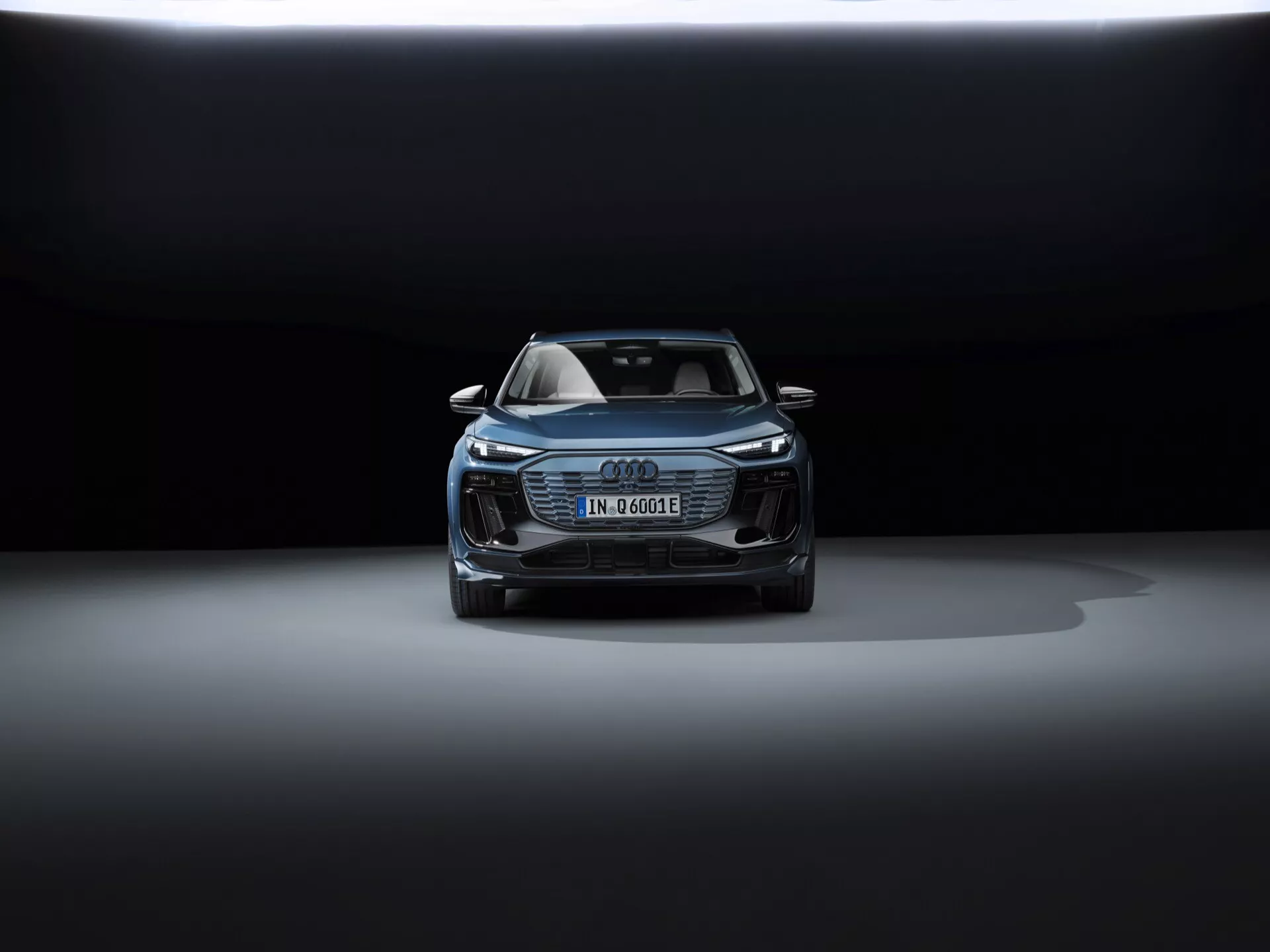 Pogled z sprednje strani na 2024 Audi Q6 e-tron, ki prikazuje pokrov motorja, rešetko, del strehe in sprednji dizajn. Elegantni žarometi verjetno uporabljajo LED tehnologijo. Nemška registrska tablica nakazuje na električni ali hibridni model. Značilna Audi Singleframe rešetka, dodatni zračni vhodi in morebitni varnostni senzorji, ki poudarjajo napredne tehnologije pomoči vozniku. Dramatična osvetlitev s temnim ozadjem in osvetlitev od zgoraj poudarjata luksuzni in prefinjeni dizajn avtomobila.