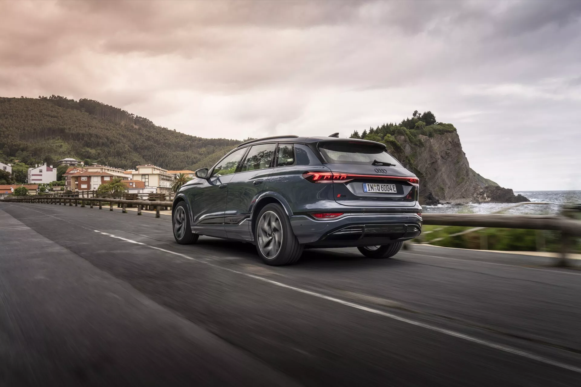 Audi Q6 e-tron 2024 posnet od zadaj iz treh četrtin med vožnjo po obalni cesti, ki prikazuje značilne LED zadnje luči, temno kovinsko barvo, lita platišča in praktičen zadnji brisalnik. Zamegljeno ozadje kaže na obalno okolje, ki odraža estetiko in tehnološki napredek sodobnega SUV-ja.